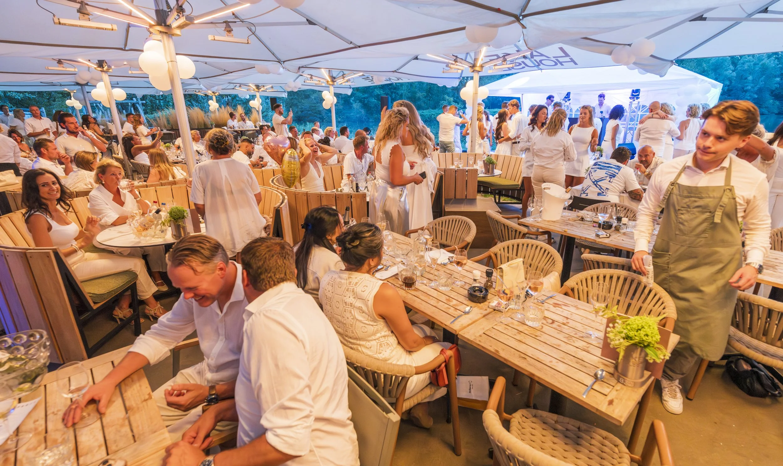 Een grote groep mensen die zich verzameld heeft op een feest of bijeenkomst onder een overkapping met witte lampionnen, met mensen die zitten aan tafels en anderen die dansen en drinken.