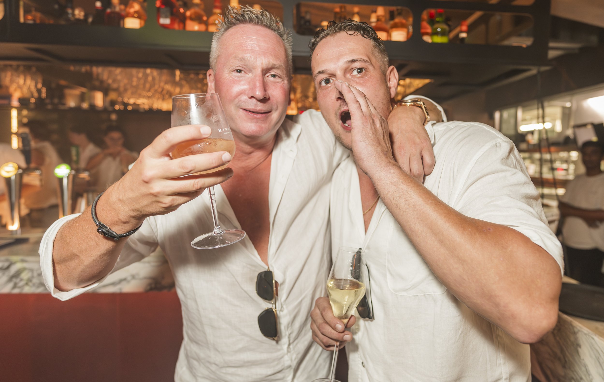 Twee mannen vieren feest in een bar, een houdt een glas wijn omhoog terwijl de ander schreeuwt en zich de hand voor de mond houdt.