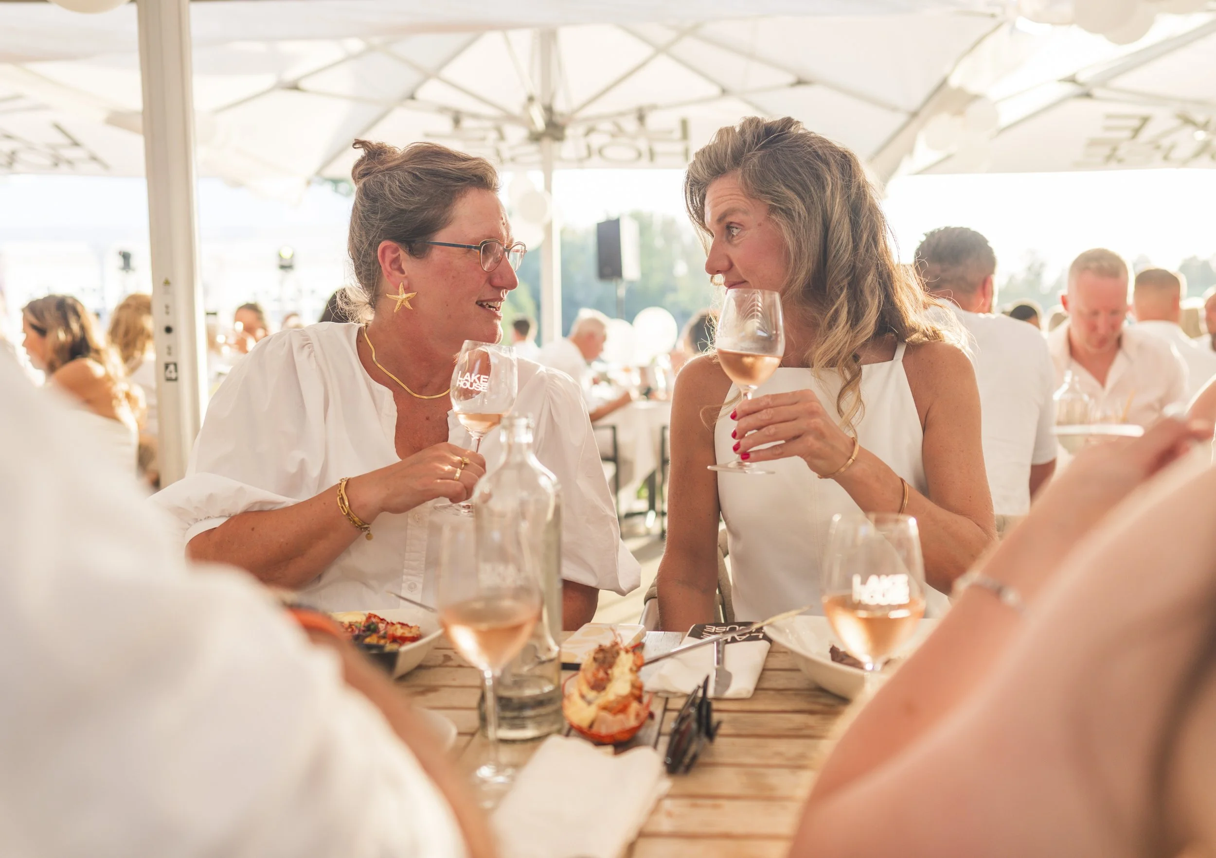 Twee vrouwen die wijn drinken en praten tijdens een feest of bijeenkomst, met andere gasten op de achtergrond.