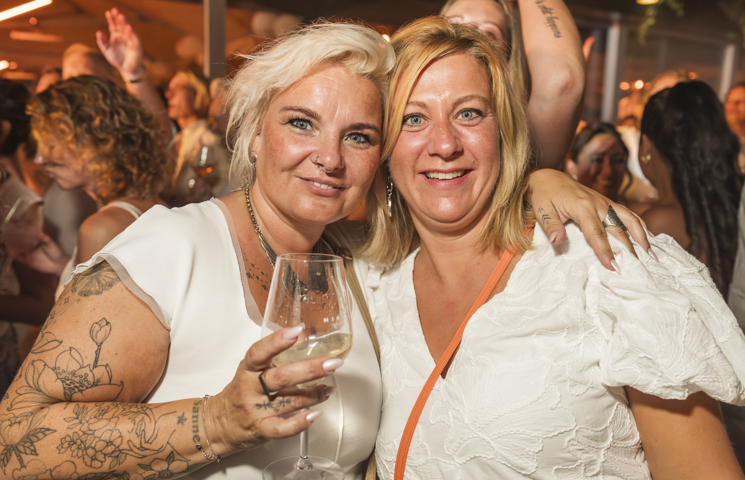 Twee vrouwen die samen in een feestelijke sfeer een glas witte wijn vasthouden, omringd door een groep mensen.