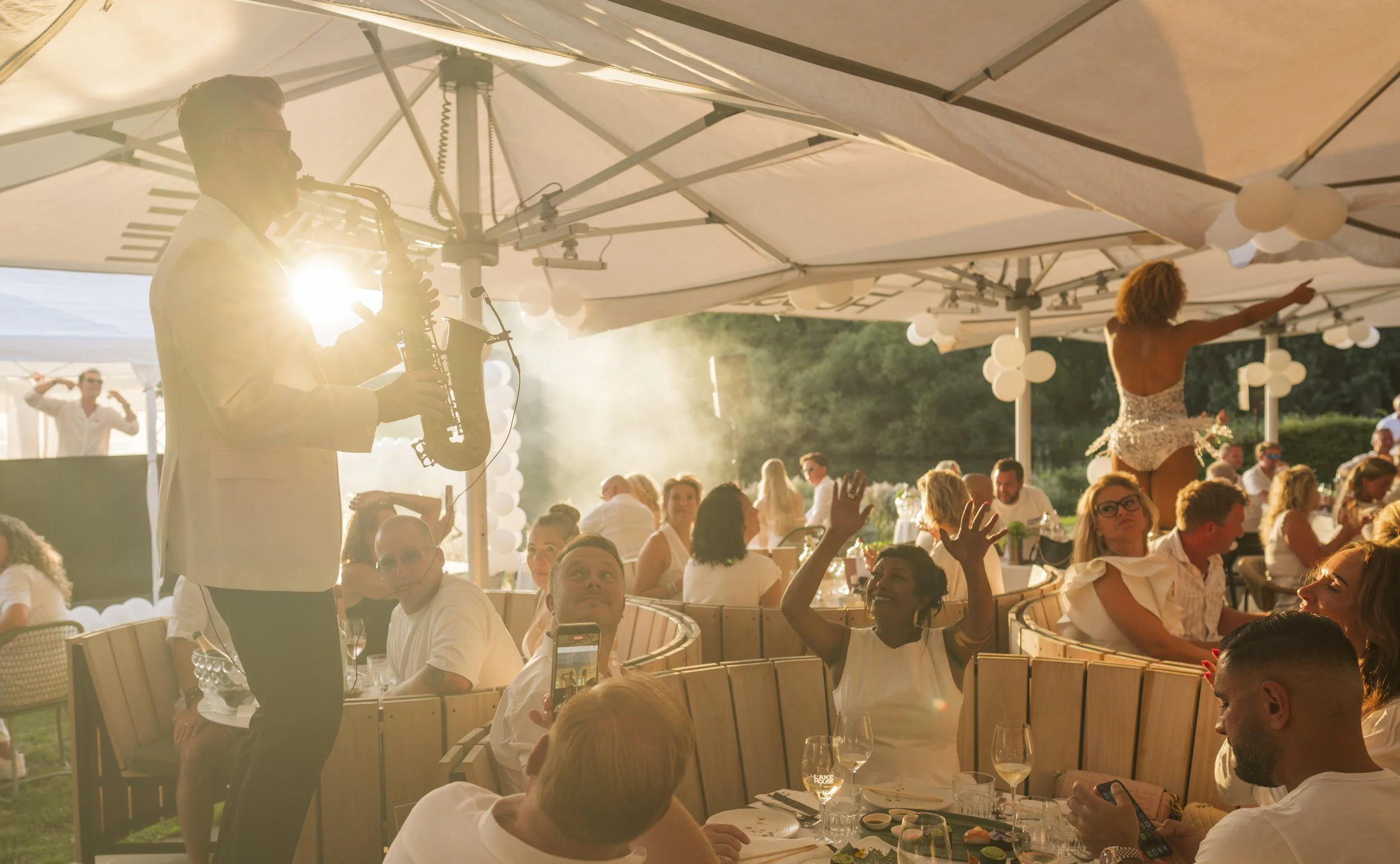 Muzikant speelt saxofoon tijdens een dinerfeest onder grote parasols met gasten die genieten en fotograferen.