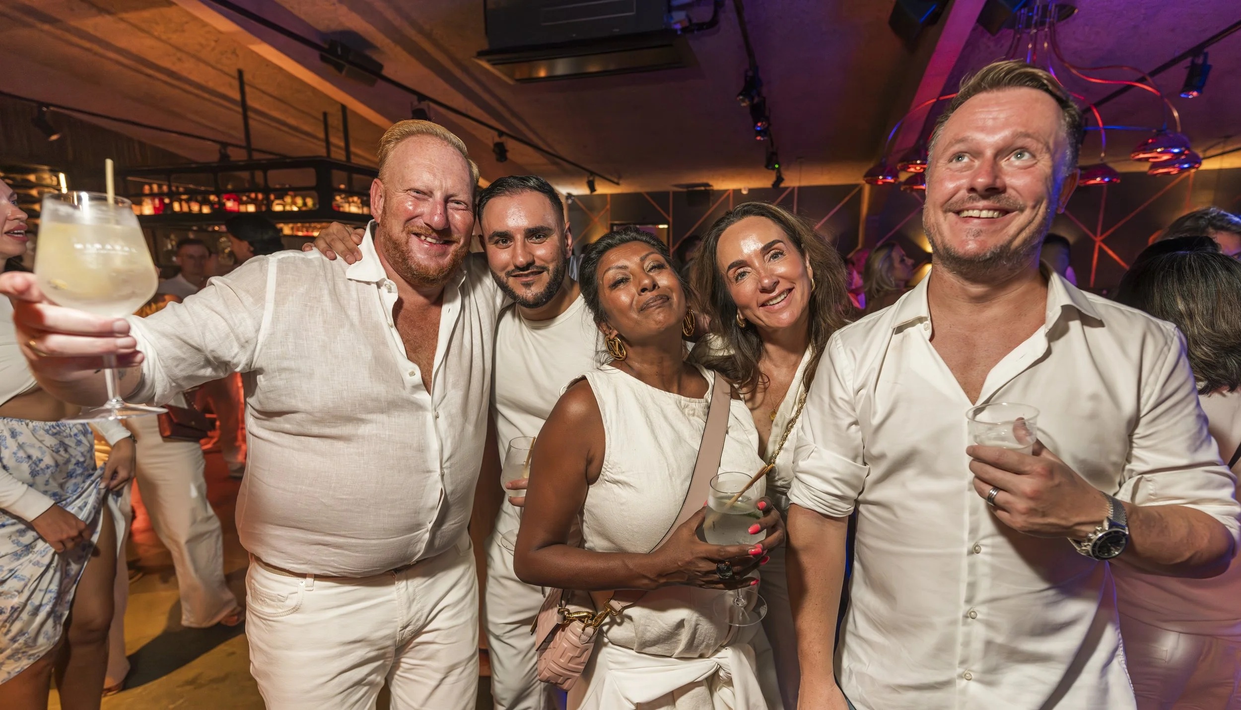 Vijf mensen op een feestje, lachend en genietend, met cocktails in hand, in een sfeervolle, kleurrijke bar of club decor.