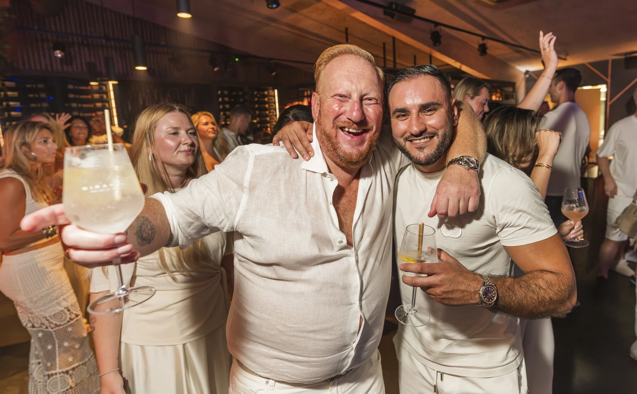Twee mannen lachen en hebben elkaar omarmd tijdens een feestje, omringd door andere feestgangers in een warme, sfeervolle locatie.