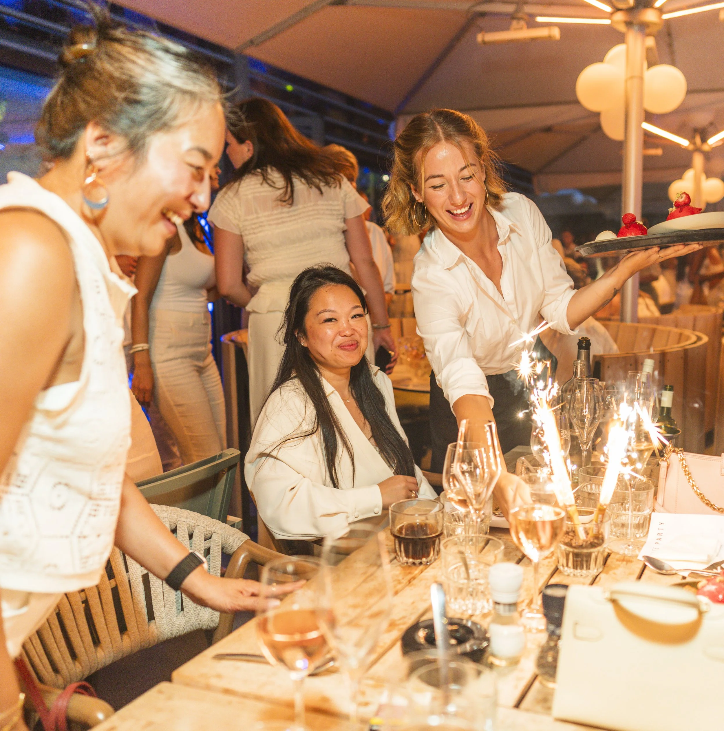 Mensen lachen en genieten bij een feestelijke gebeurtenis, met wijn en desserts op tafel, onder warme lichtinrichting.