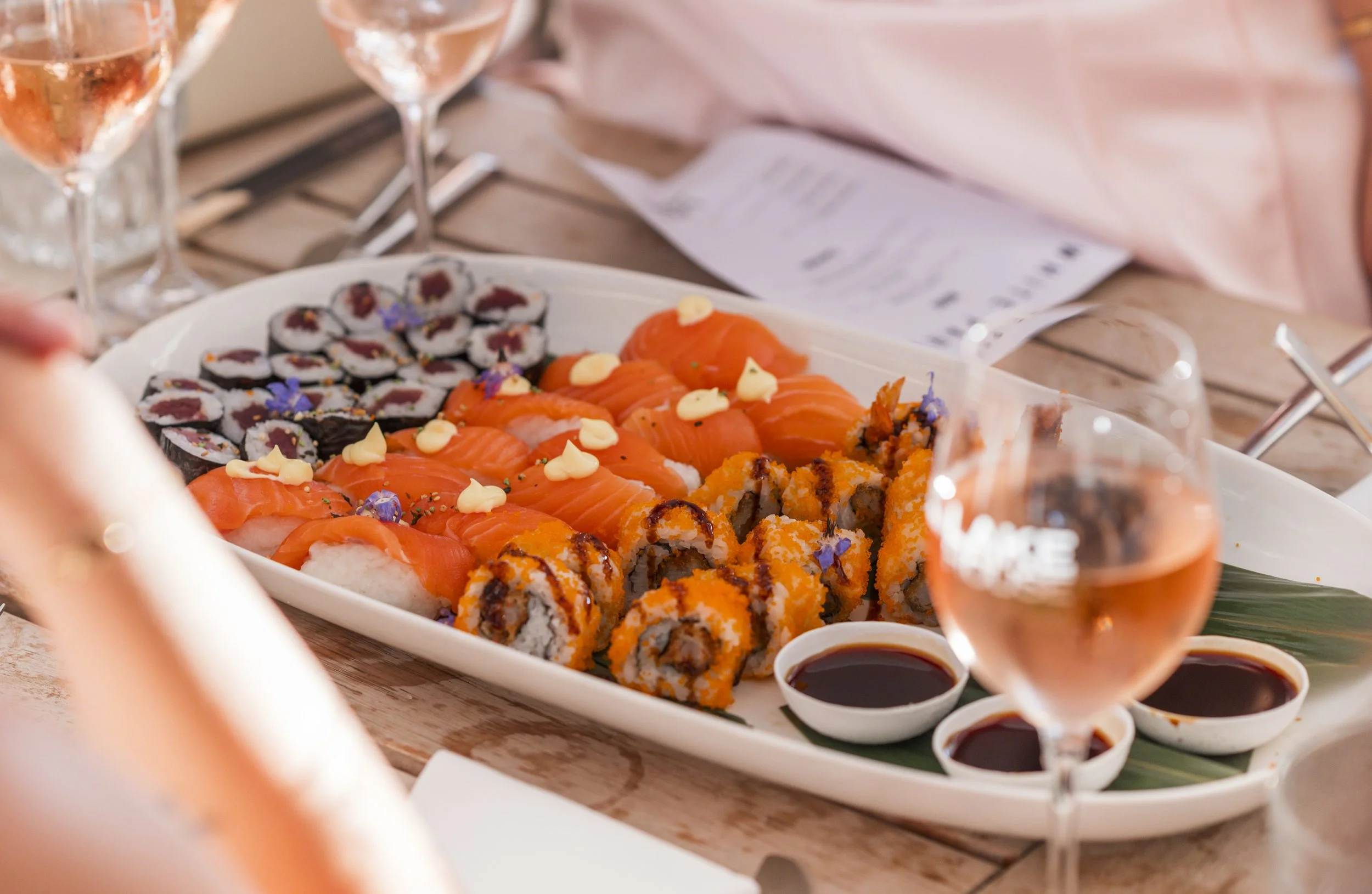 Sushi schaal met verschillende soorten sushi, roze wijn in glazen, sojasaus in kleine schaaltjes, en een verslag van een eetmoment op een houten tafel.