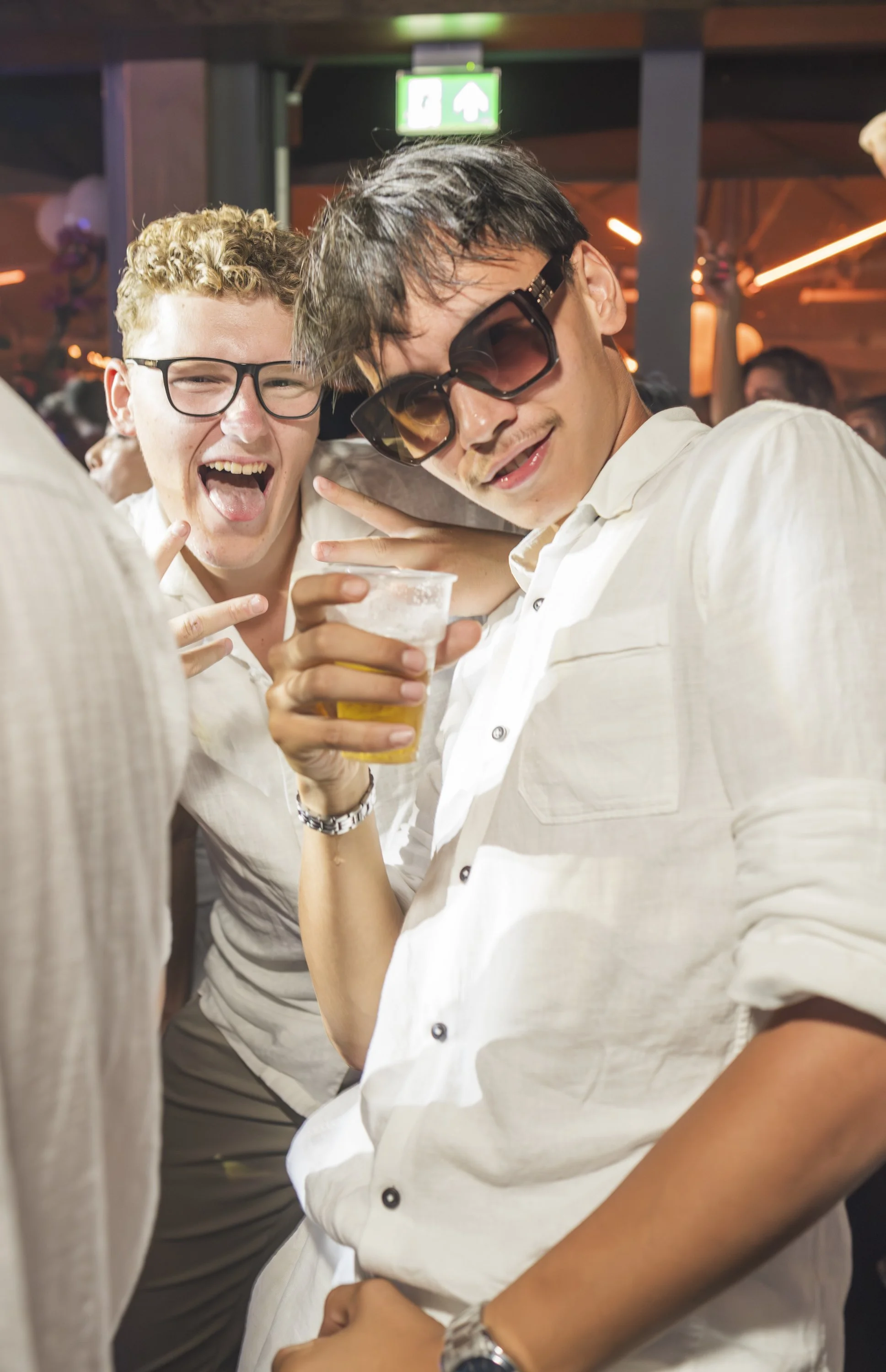 Twee jonge mannen die plezier maken op een feestje, één met een glas bier en de ander met een peace-teken, in een drukke feesttent met gekleurd licht.
