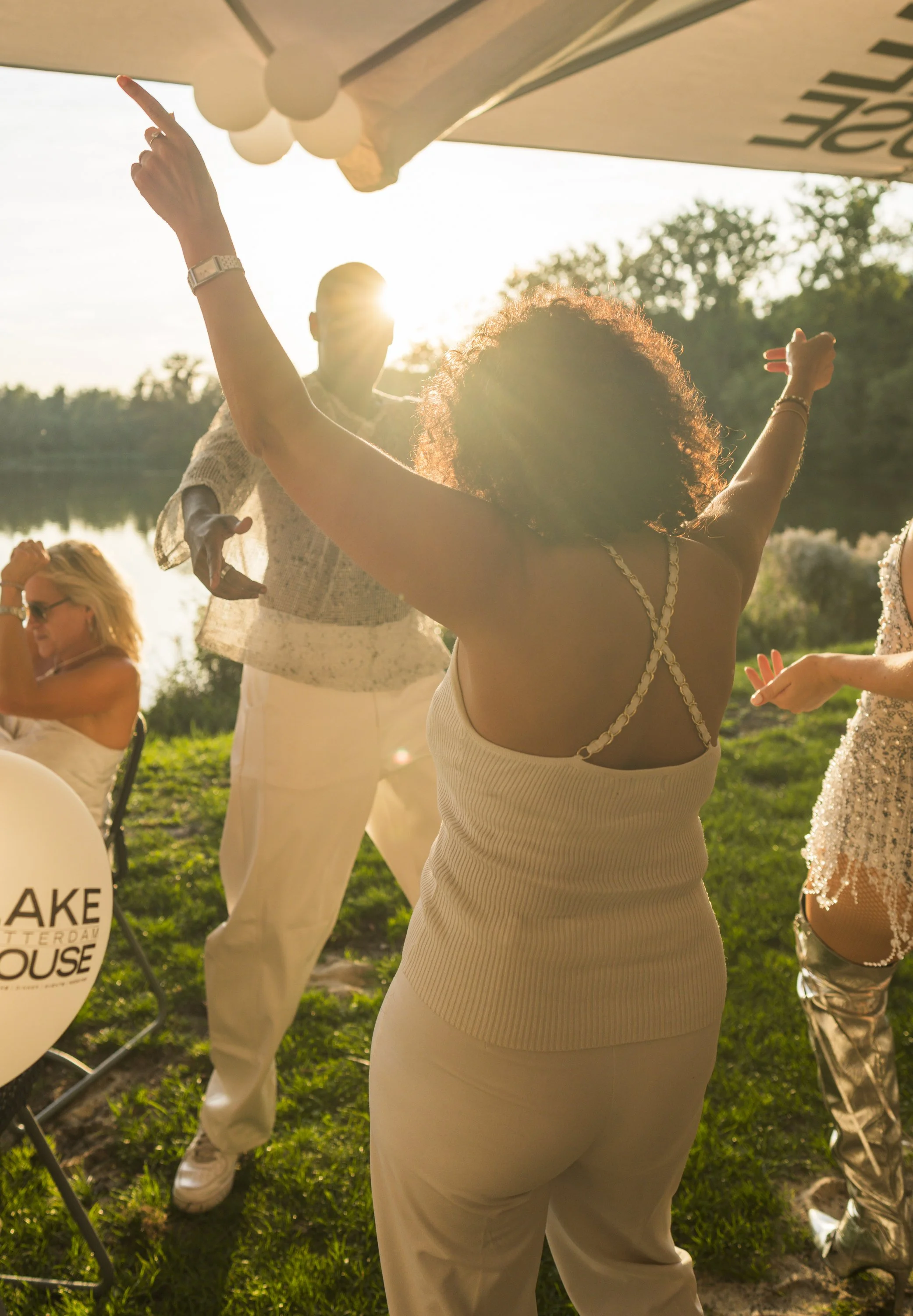 Mensen dansen buiten in de zon, vrouw met gebogen armen en dansbewegingen, achtergrond van water en bomen, feestelijke sfeer.