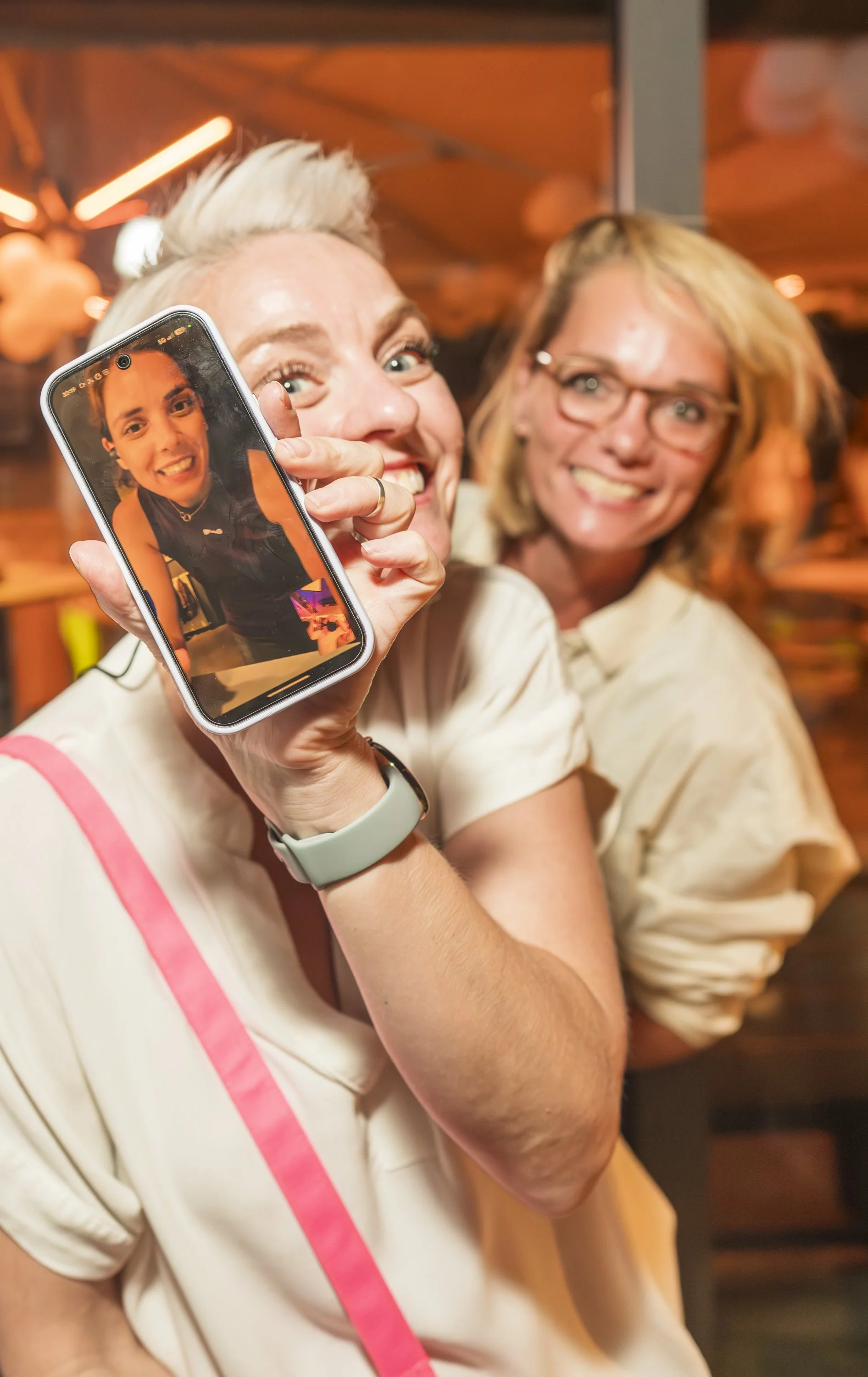 Twee vrouwen lachen terwijl een vrouw een foto maakt met haar smartphone in een gezellige, warme binnenruimte.