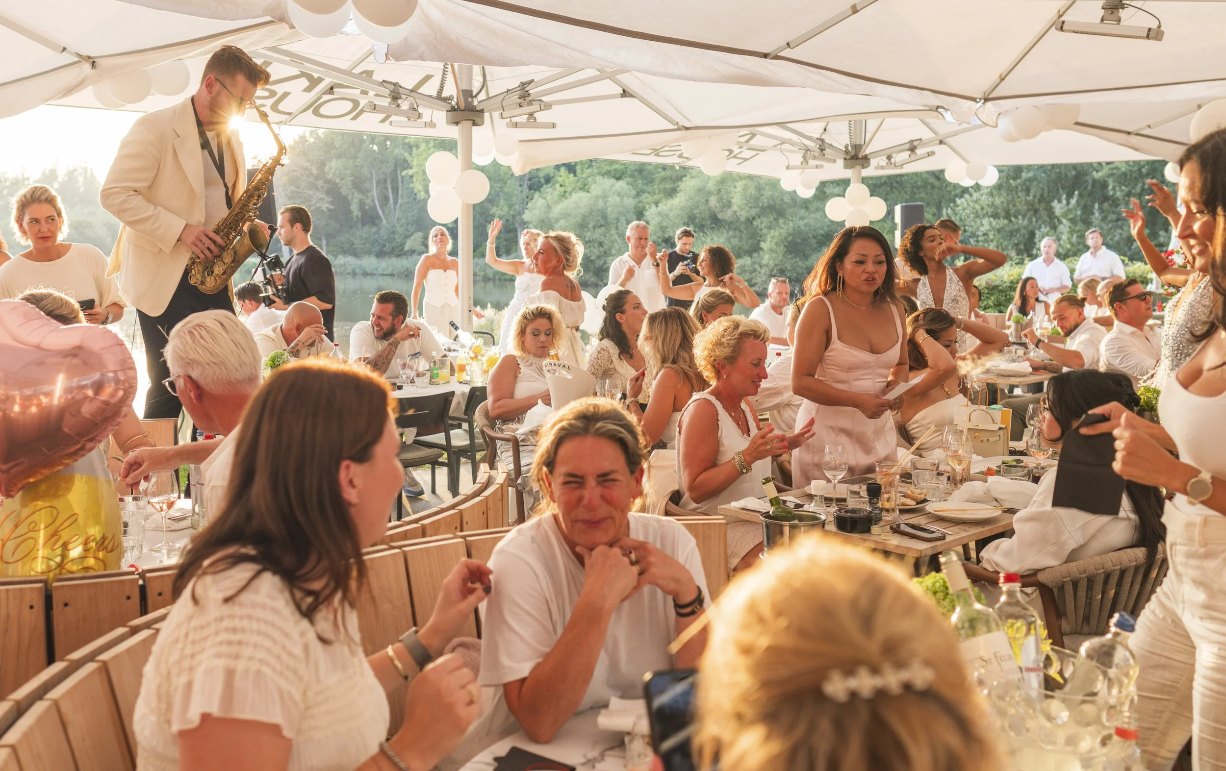 Een groep mensen die genieten van een buitenbrunch onder grote parasols met witte lampionnen, terwijl een muzikant saxofonist optreedt.