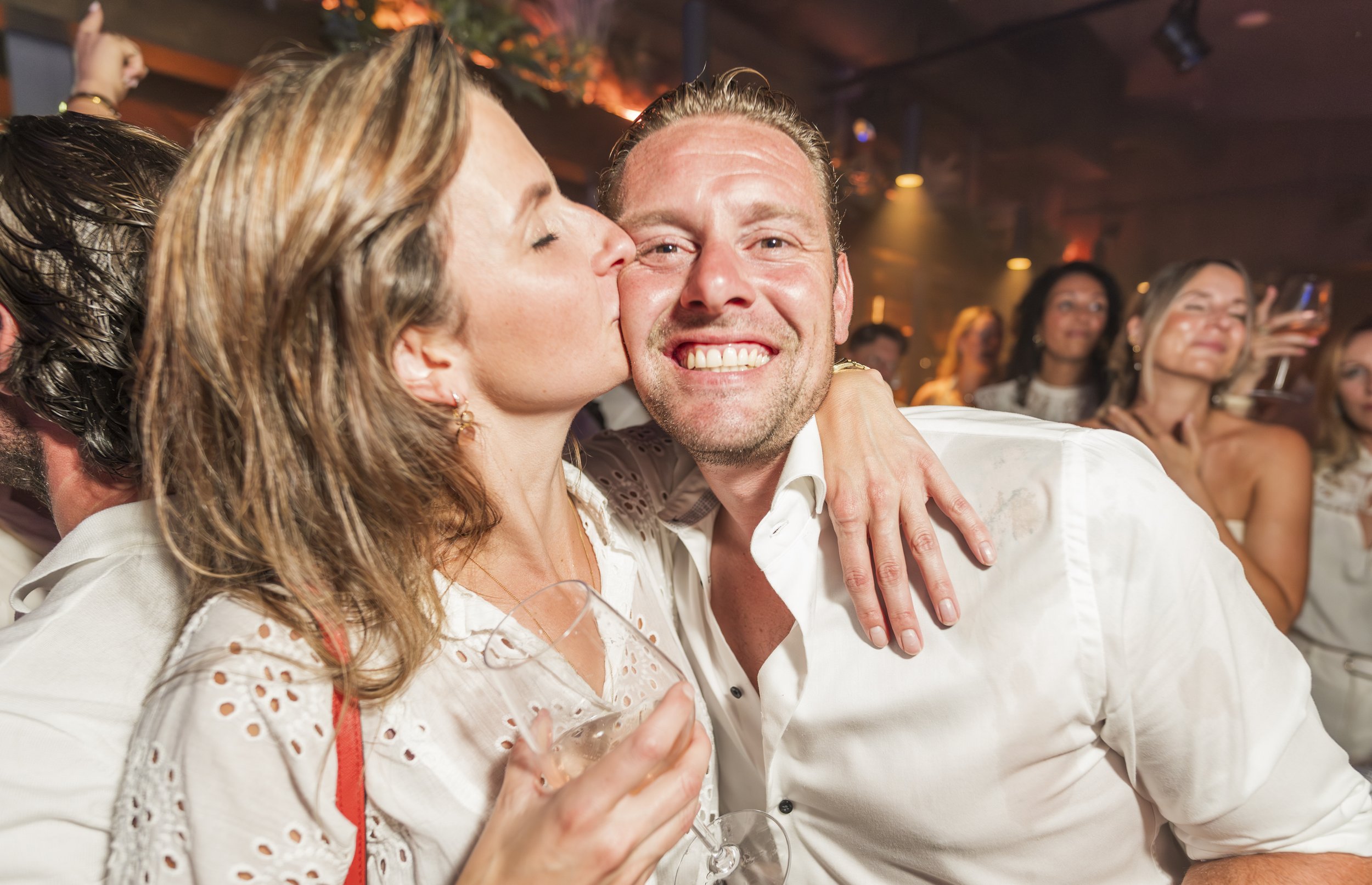Een man en een vrouw knuffelen en lijken te genieten op een feestje. De vrouw kust de man op de wang, terwijl hij lacht. Op de achtergrond is een groep mensen te zien die meedoen aan het feest.