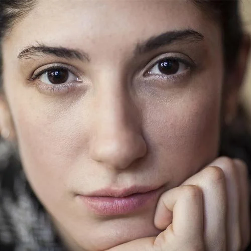 Primer plano de rostro de joven mujer con expresión pensativa, mirando hacia la cámara, con la mano tocando su mentón.