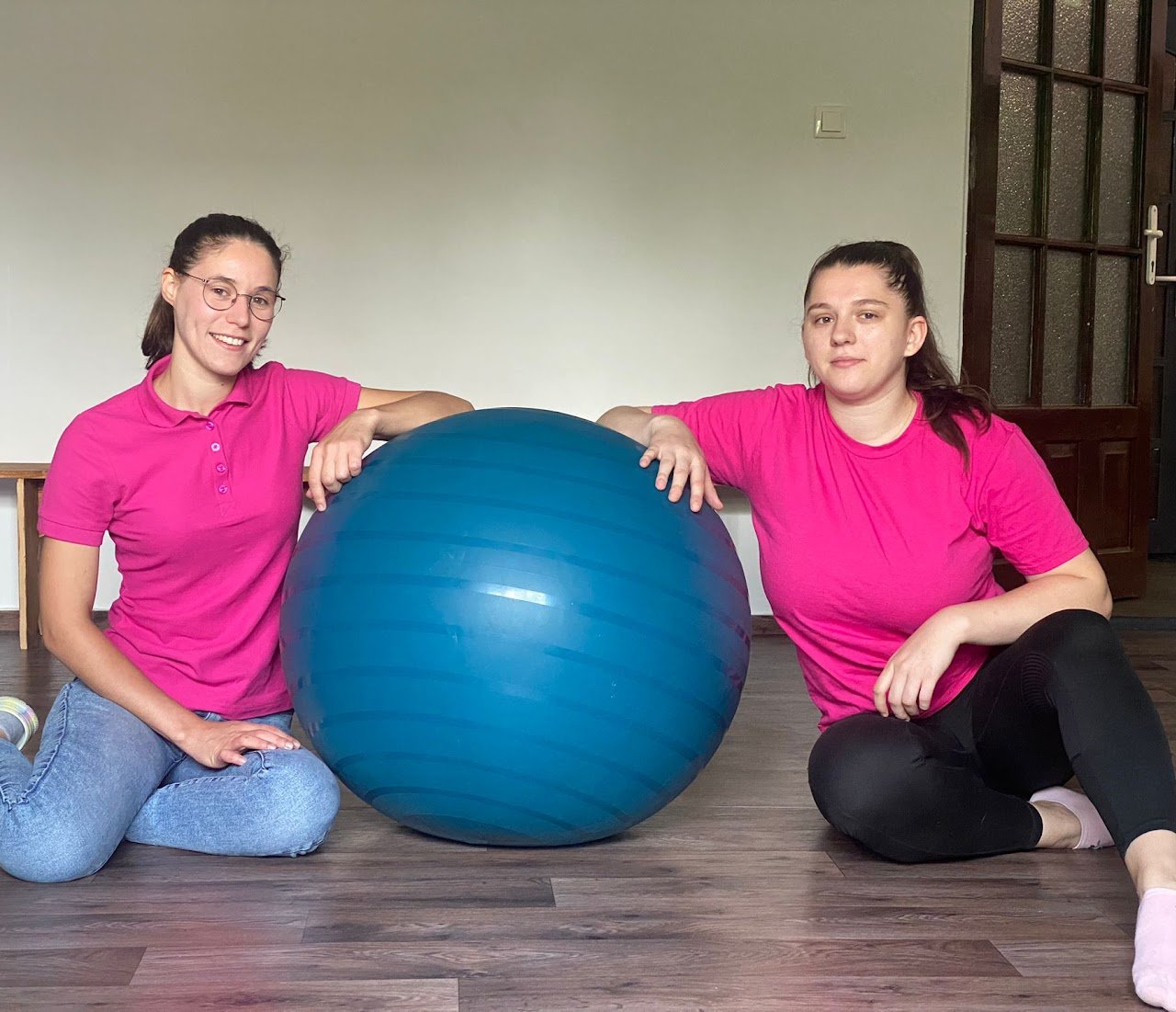 Two women in pink shirts sitting on a wooden floor with a large blue exercise ball between them, smiling and resting their hands on the ball.