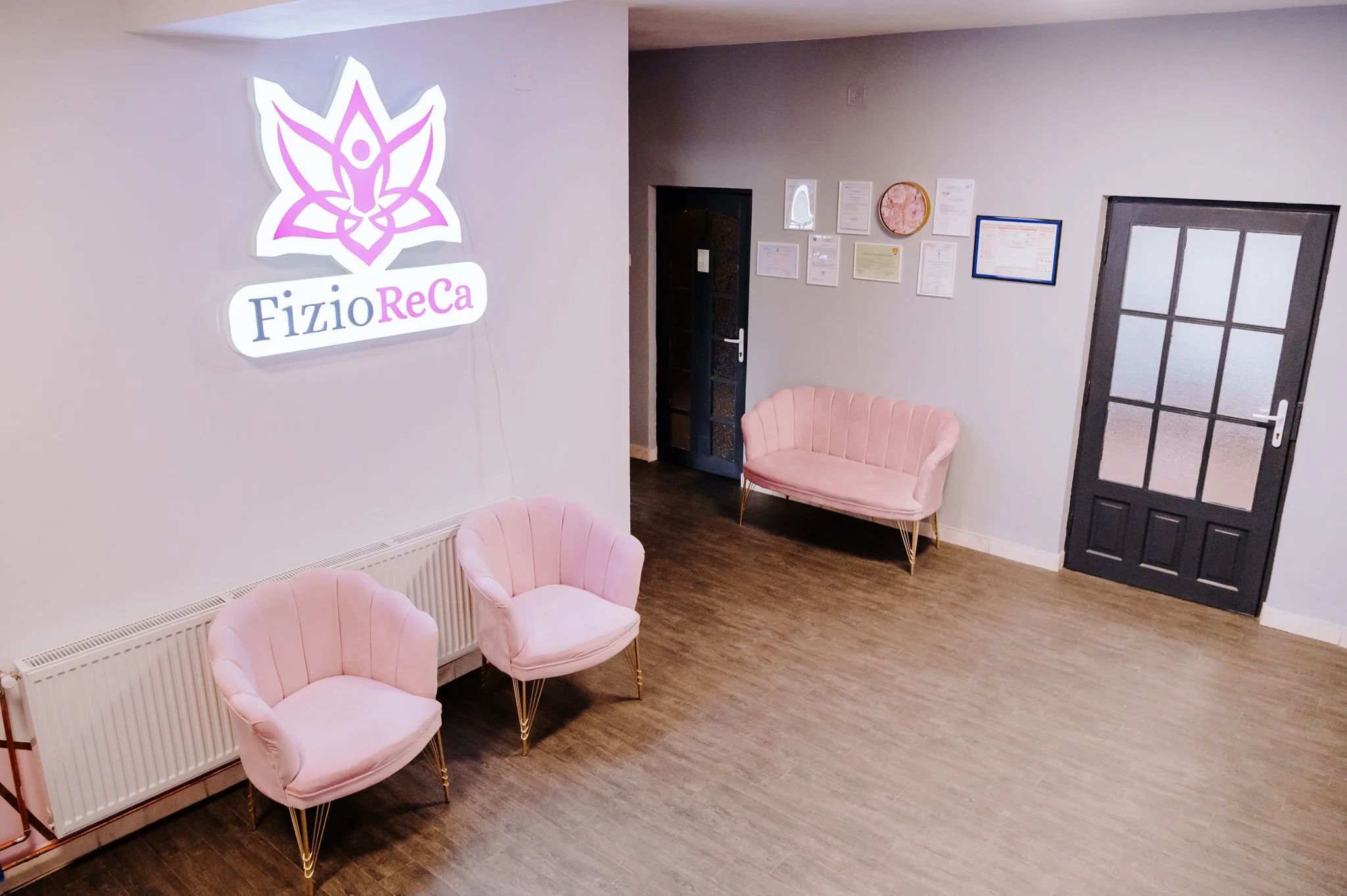 Waiting room with three pink velvet chairs, a wall with certificates and a decorative framed piece, and a bright logo sign that reads 'FizioReCa' on the wall.