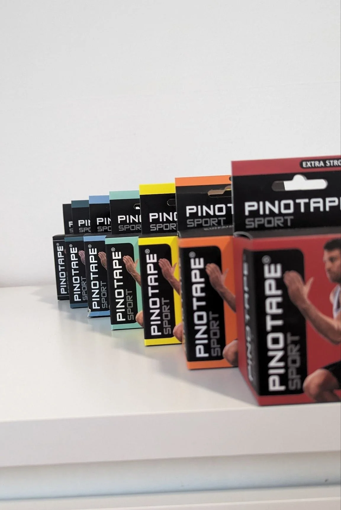 A line of various colored boxes of PINOTAPE SPORT athletic tape on a white surface, with the boxes arranged from left to right in blue, light blue, green, yellow, orange, and red colors.