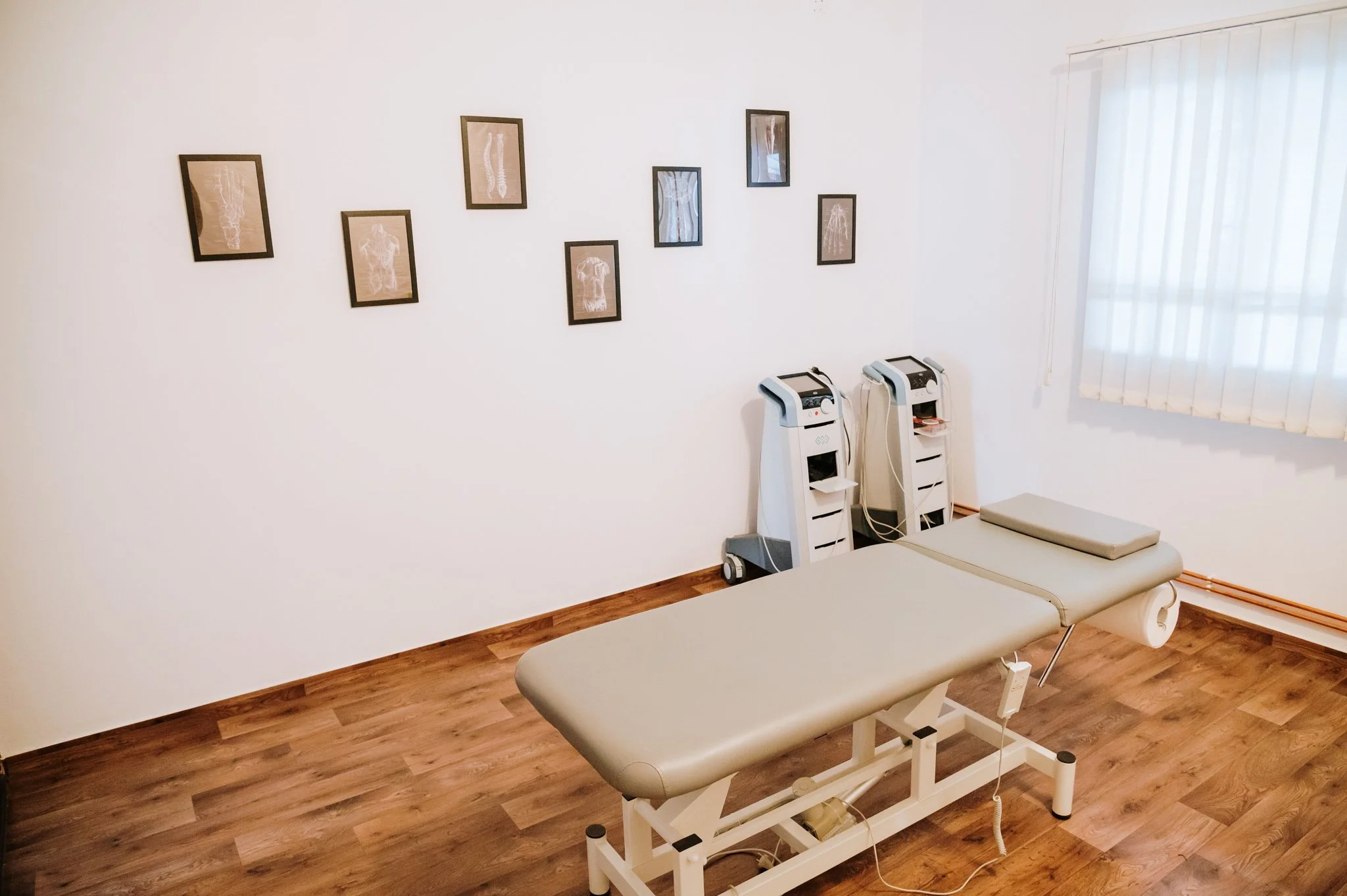 Empty medical examination room with a beige treatment table, two medical machines, framed x-ray images on the wall, and a window with blinds.
