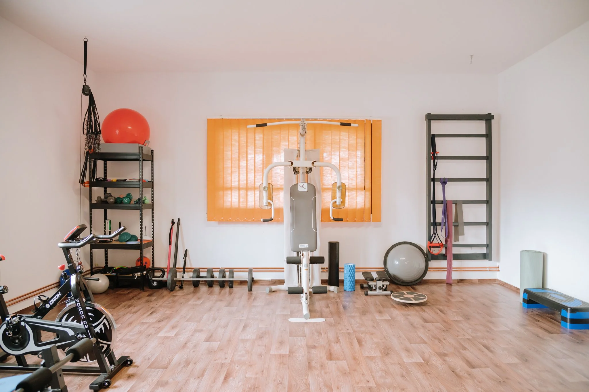 Home gym with exercise equipment, weights, stability ball, resistance bands, step platform, and a wall-mounted ladder for workouts, with orange curtains covering the window.
