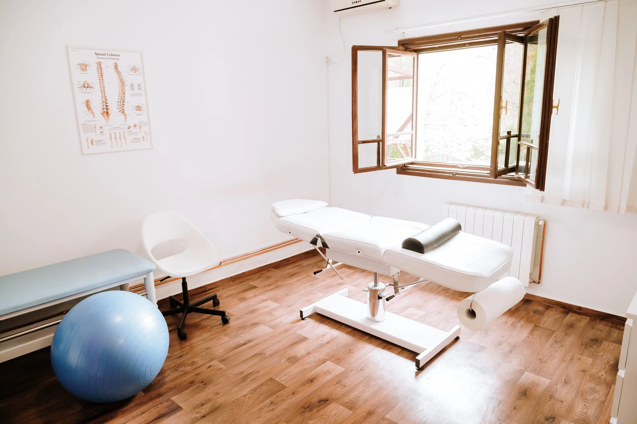 Medical examination room with a treatment table, an exercise ball, a white chair, a small bench, and a window with open wooden shutters, wooden flooring, and a spine anatomy poster on the wall.