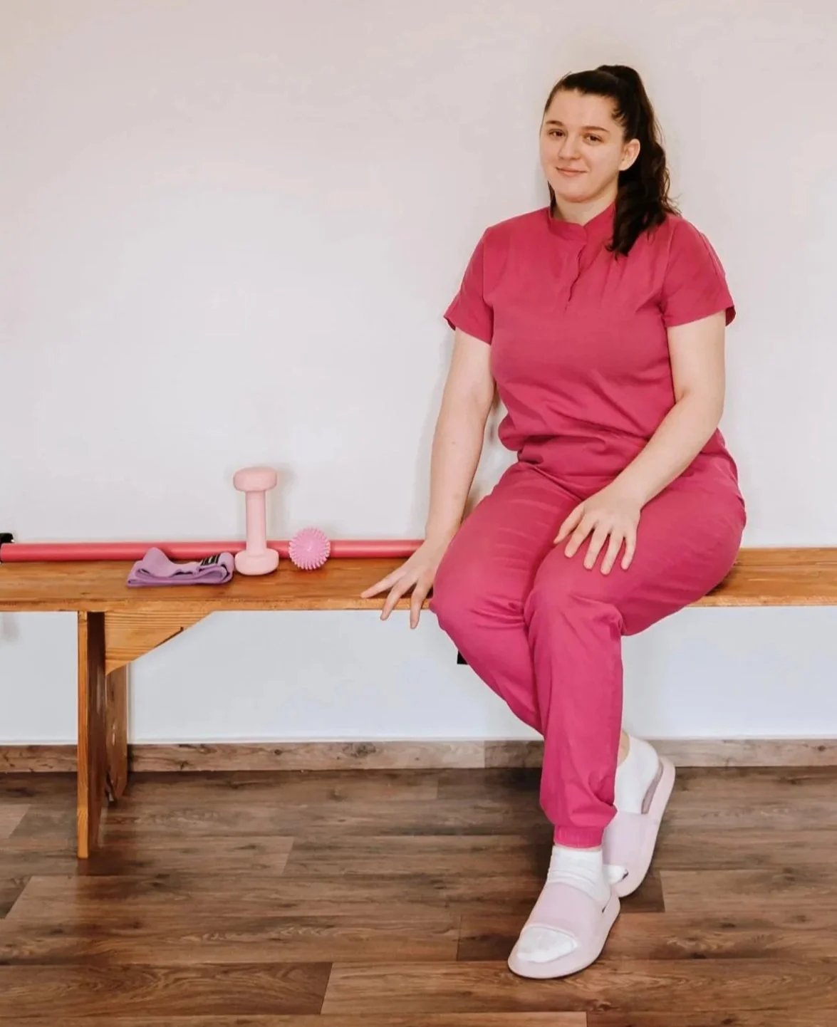 A woman in pink scrubs sitting on a wooden bench with small fitness equipment next to her, including a pink dumbbell, pink massage ball, and purple towel.
