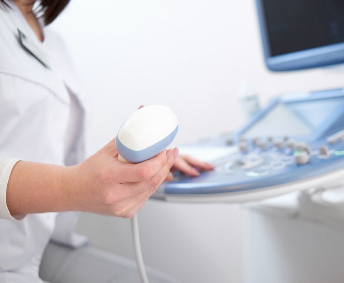 Person holding an ultrasound transducer in a medical setting with ultrasound machine in the background.