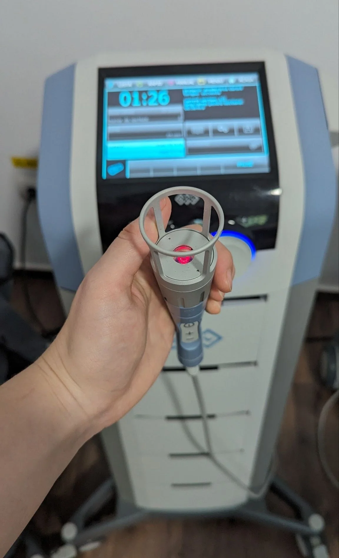 A person holding a medical device with a red laser, pointed at an advanced medical machine with a digital screen, in a clinical setting.