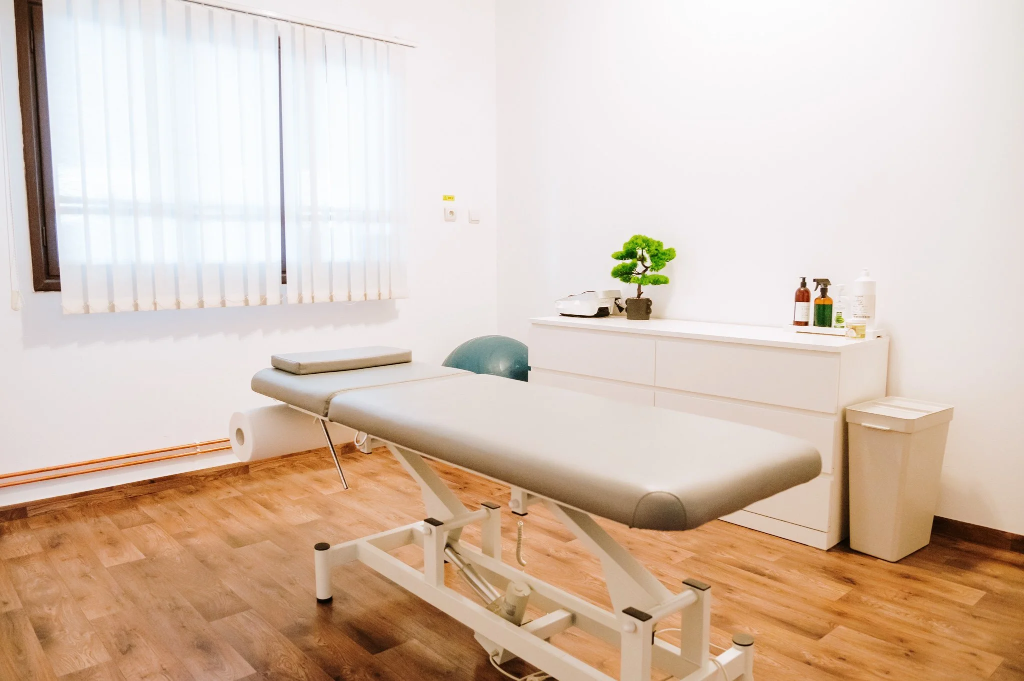 An empty medical examination room with a massage or examination table, a window with blinds, a white cabinet with bottles and a small plant, and a trash bin.