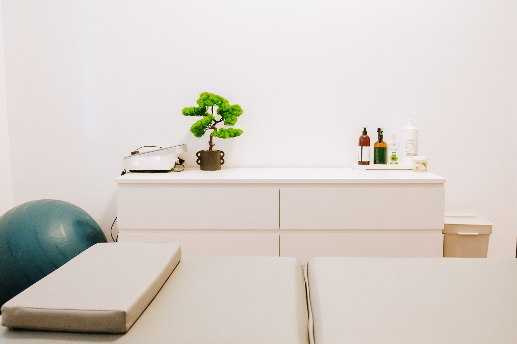 Medical treatment room with a treatment table, a small cushion, and a blue exercise ball. A white cabinet behind holds a potted bonsai tree, a steamer, and various bottles of ointments and creams against a white wall.