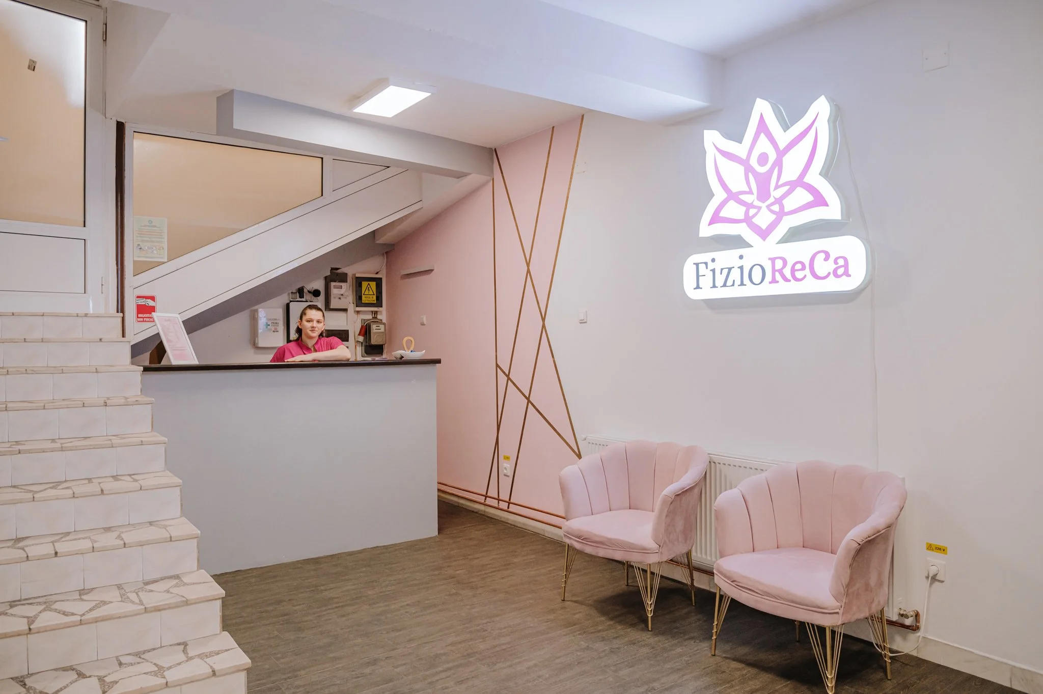 Reception area with two pink chairs, a woman at the counter, neon sign reading 'FiziorEca' with a lotus flower logo, stairs, and wall decor.