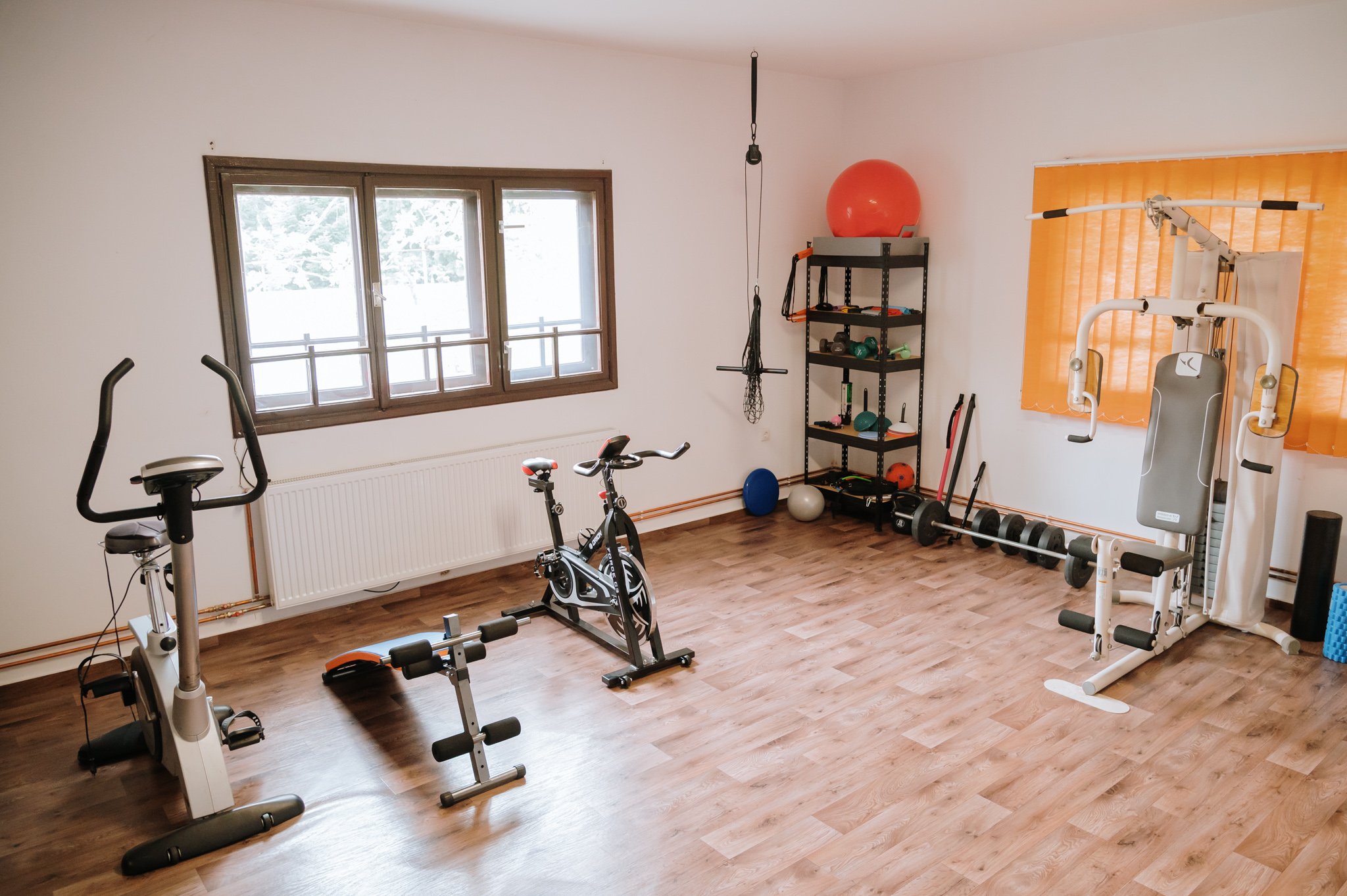 Home gym with exercise bike, elliptical, weight bench, dumbbells, and shelving with fitness equipment, wooden floor, and window with blinds.