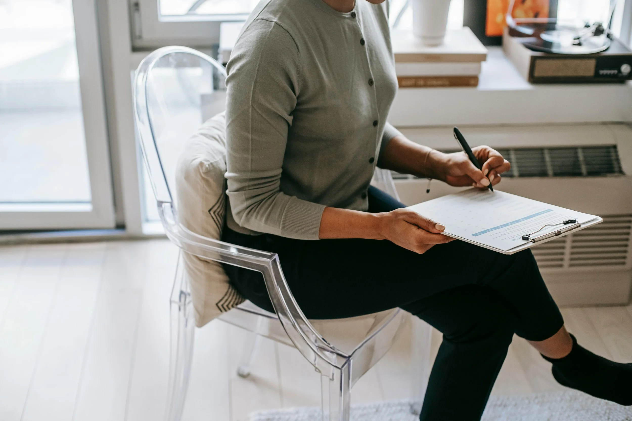Person sitting on a transparent chair, holding a clipboard and pen, working inside near a window with books and vintage record player.
