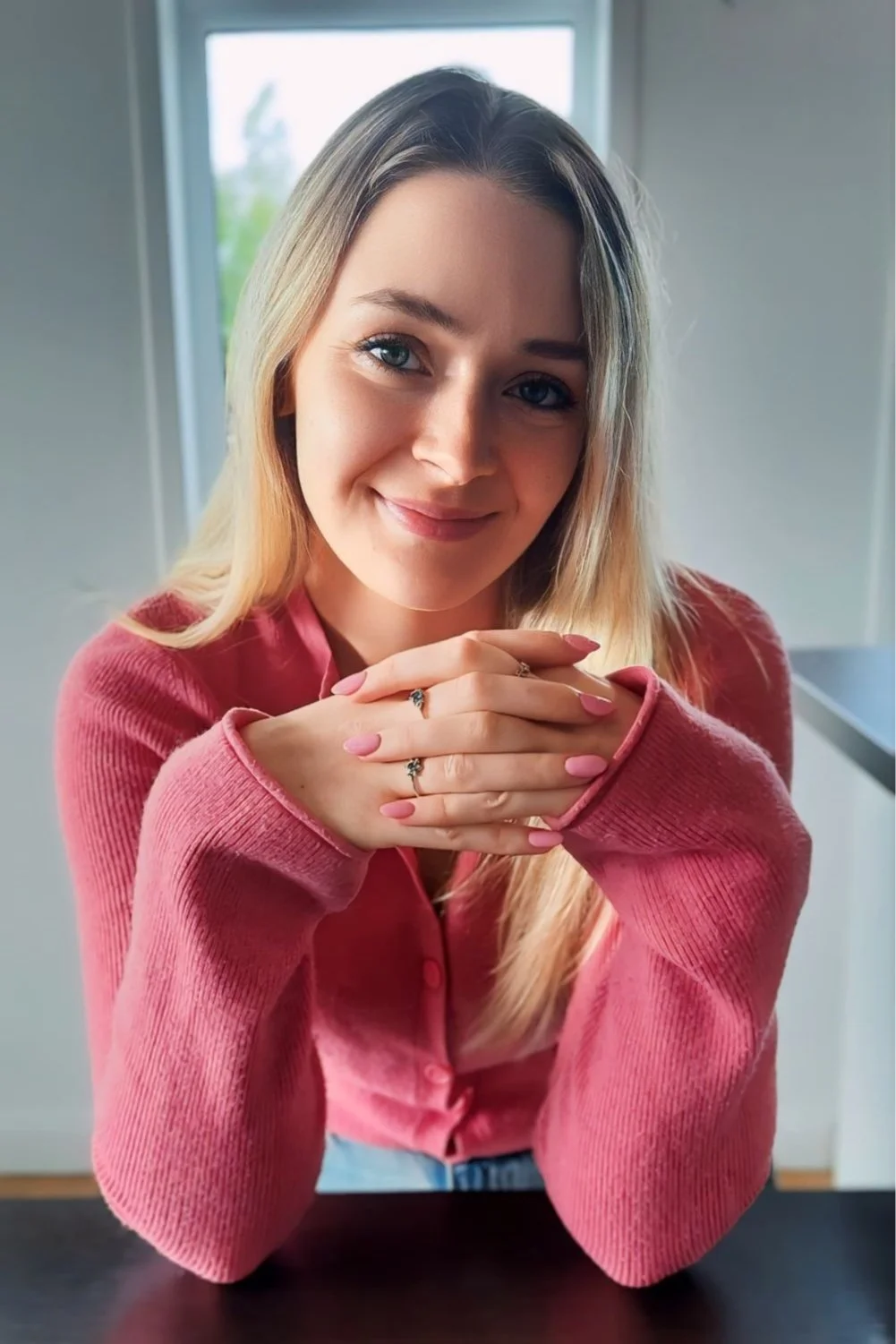 A woman with blonde hair, wearing a pink jumper, smiling and clasping her hands together, sitting indoors near a window.