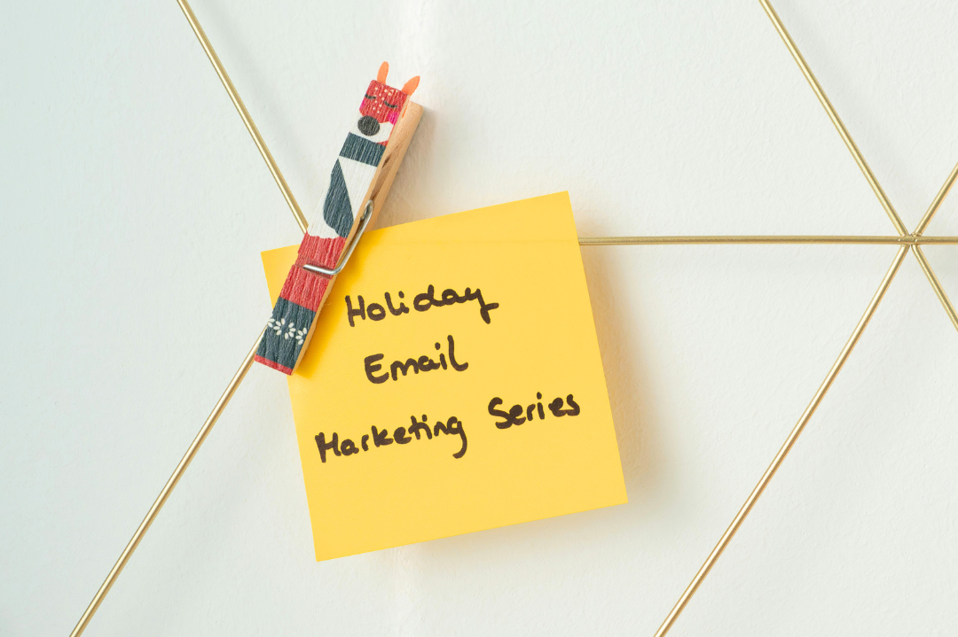 A yellow sticky note with the handwritten text 'Holiday Email Marketing Series' pinned to a white wall with a gold wire grid and a decorated clothespin featuring a reindeer or animal face.