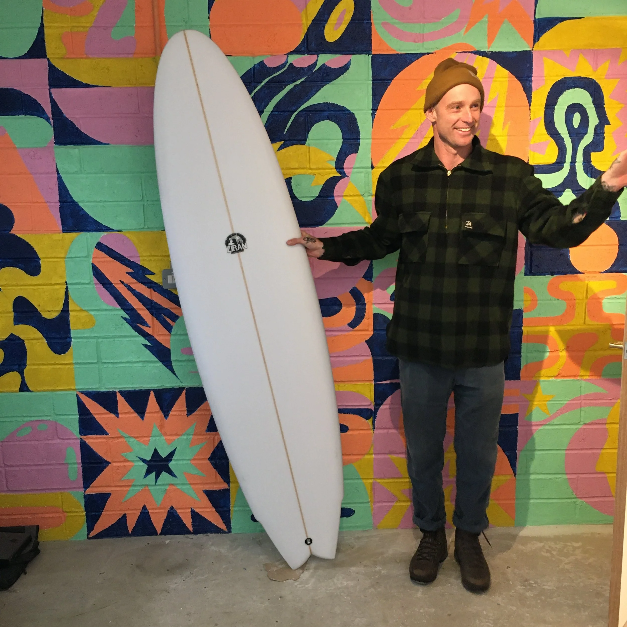 A man holding a surfboard standing in front of a colorful mural on a brick wall.