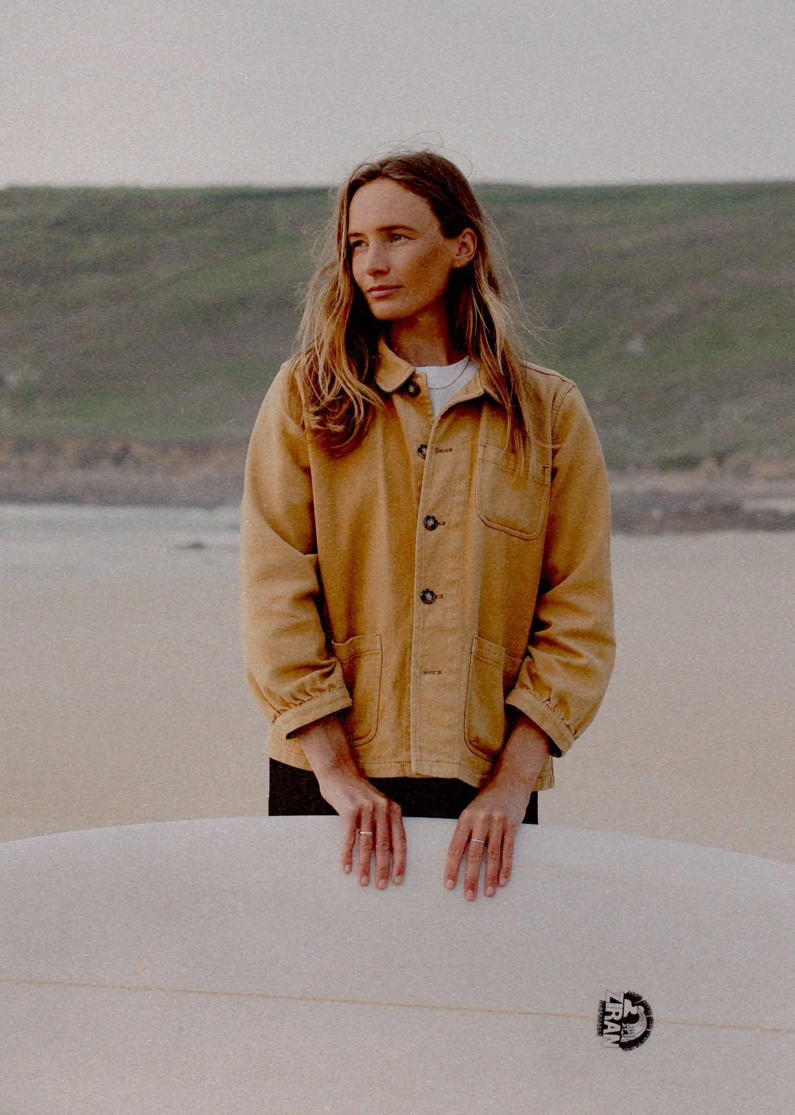 A woman standing outdoors near the beach, holding a surfboard,