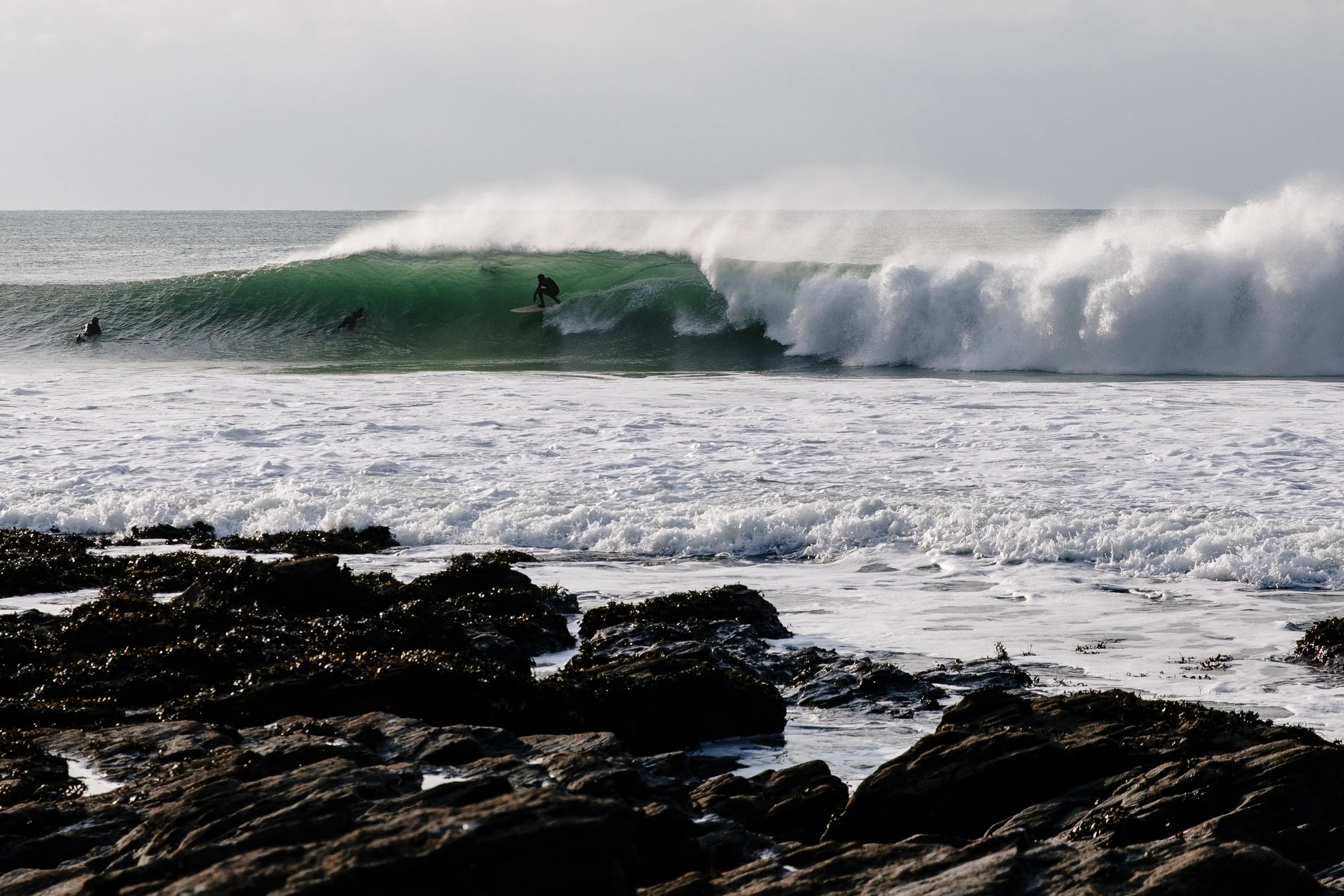 Surfers riding a large green wave near rocky shoreline. barrel barrel riding cornwall