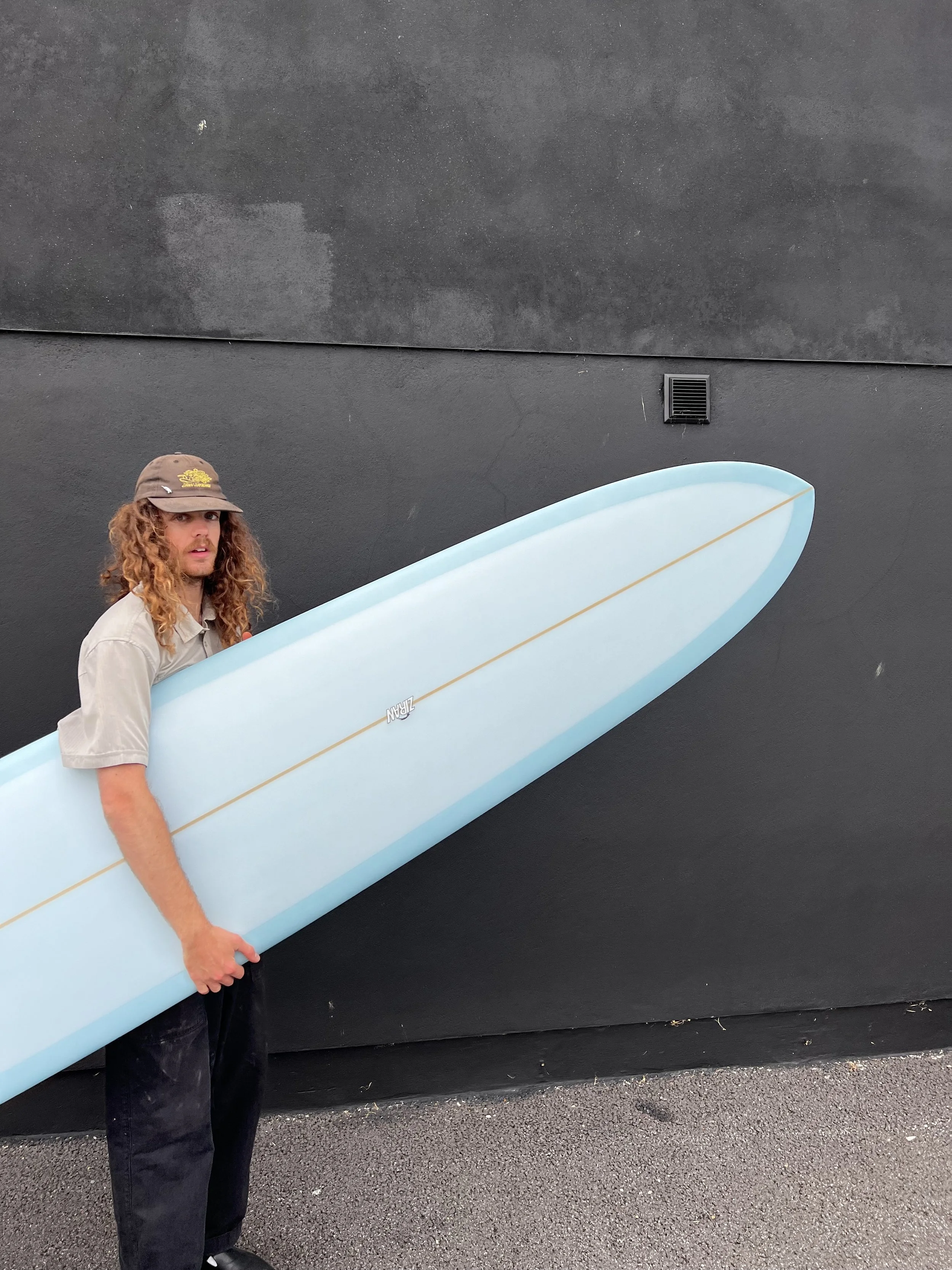 A man with long curly hair is holding a light blue surfboard against a black wall.