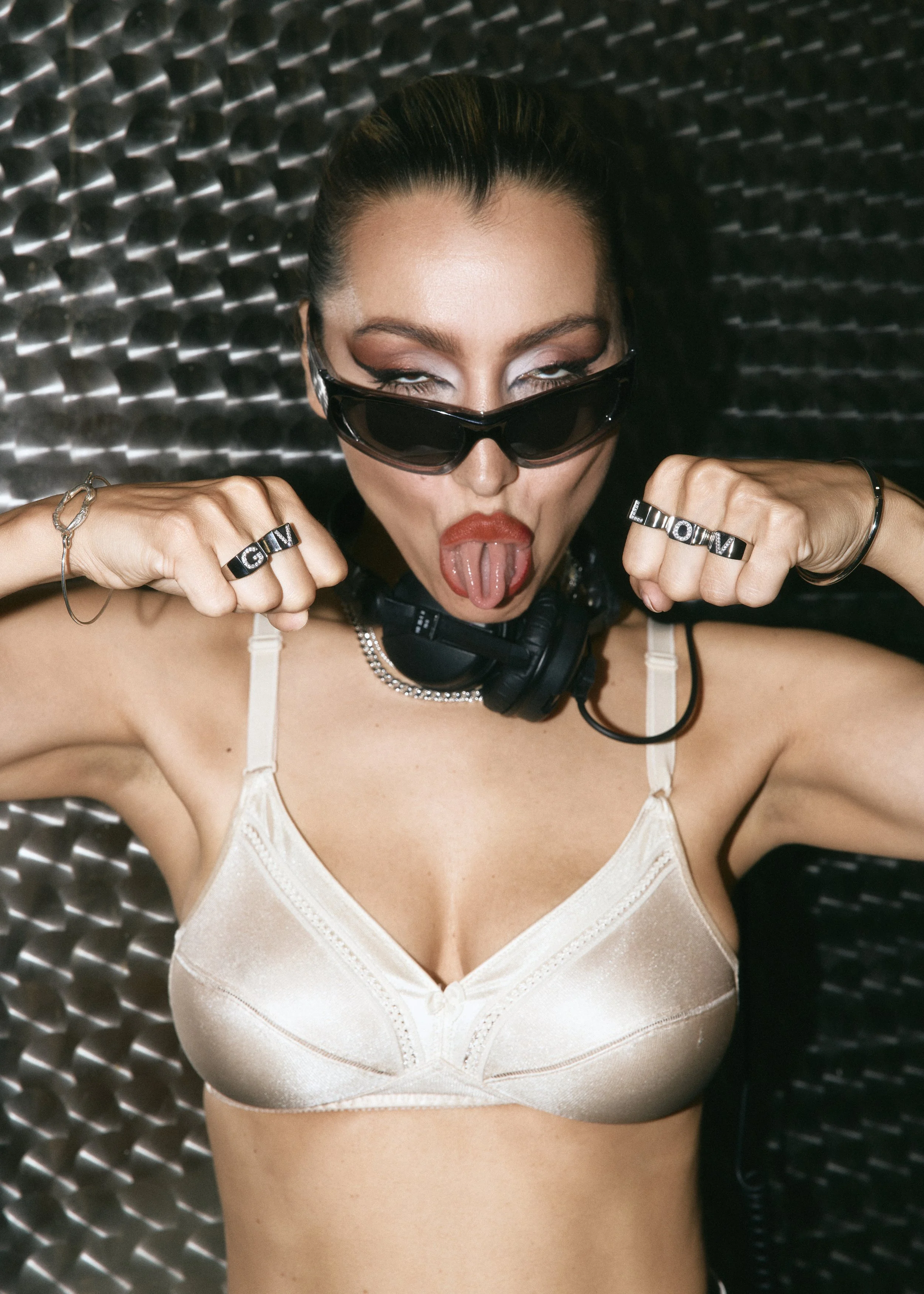 A woman with dark hair in a bun, wearing black sunglasses and rings, sticking out her tongue. She has on a cream-colored satin top and a necklace. She is lying on a textured metallic surface.