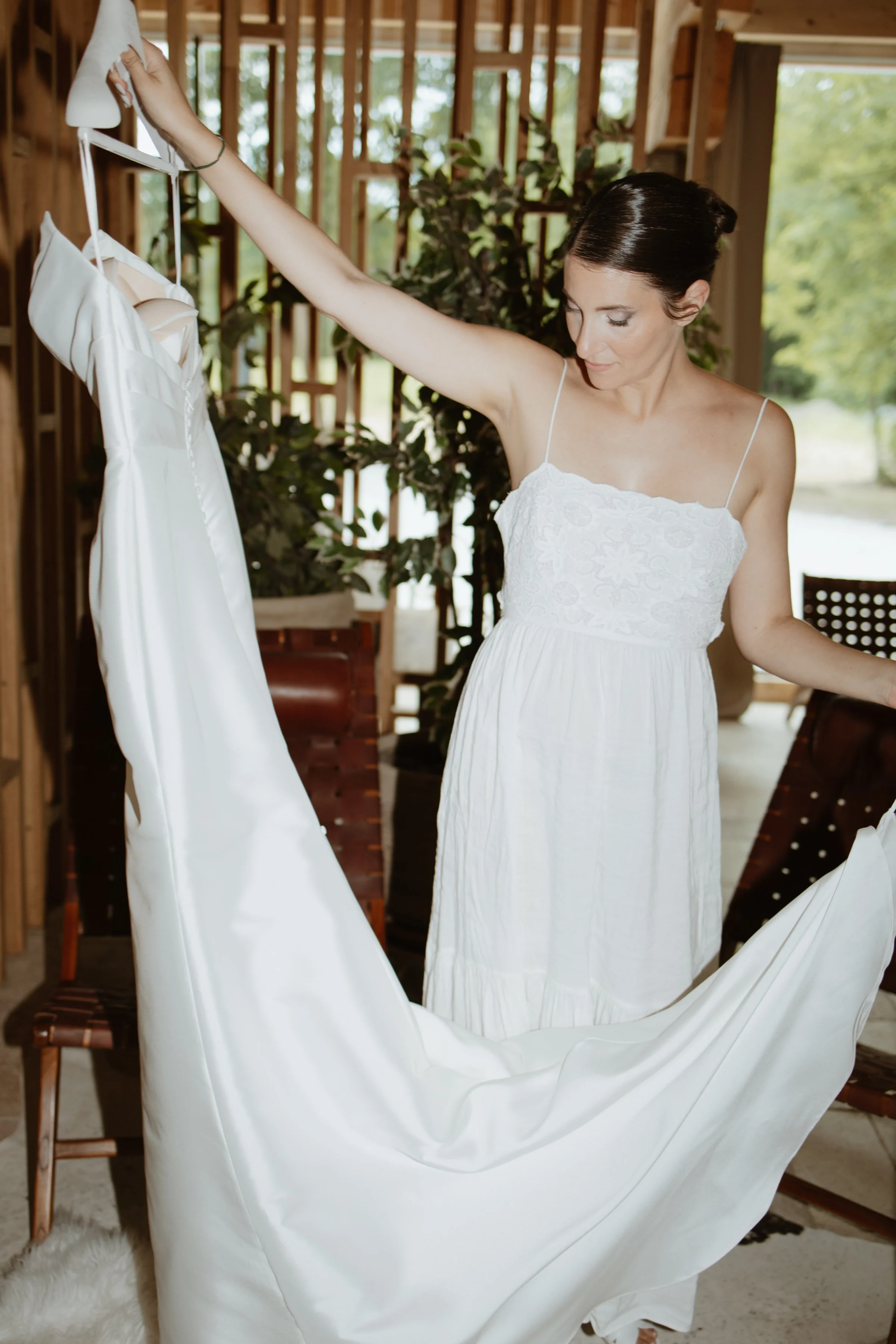 A woman with short dark hair, wearing a white dress with thin straps, is hanging a white wedding dress on a hanger in a rustic wooden room with large windows and green foliage outside.