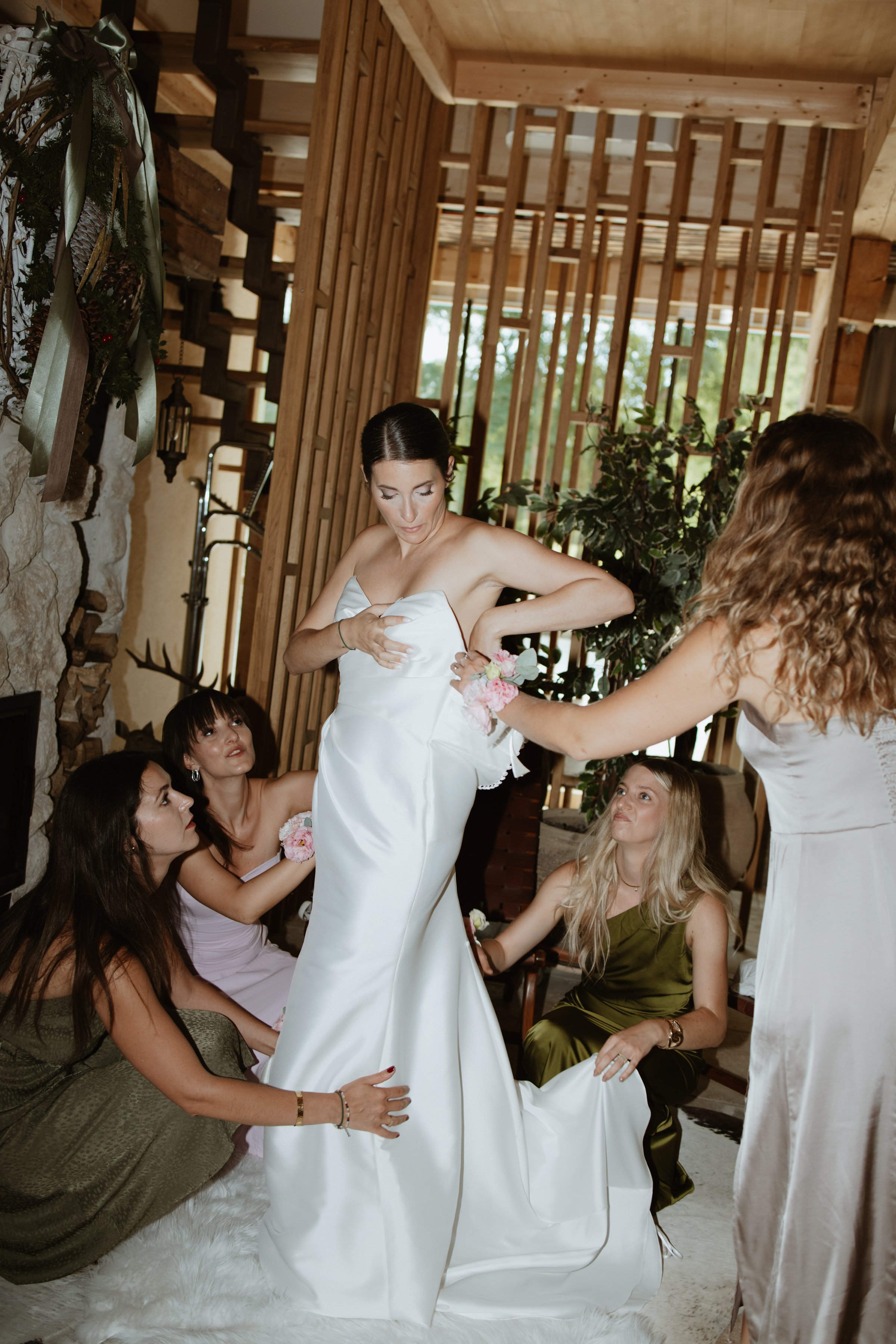A bride in a white wedding dress is sitting down, surrounded by friends who are helping her prepare. The setting is cozy with wooden walls, plants, and decorative elements.