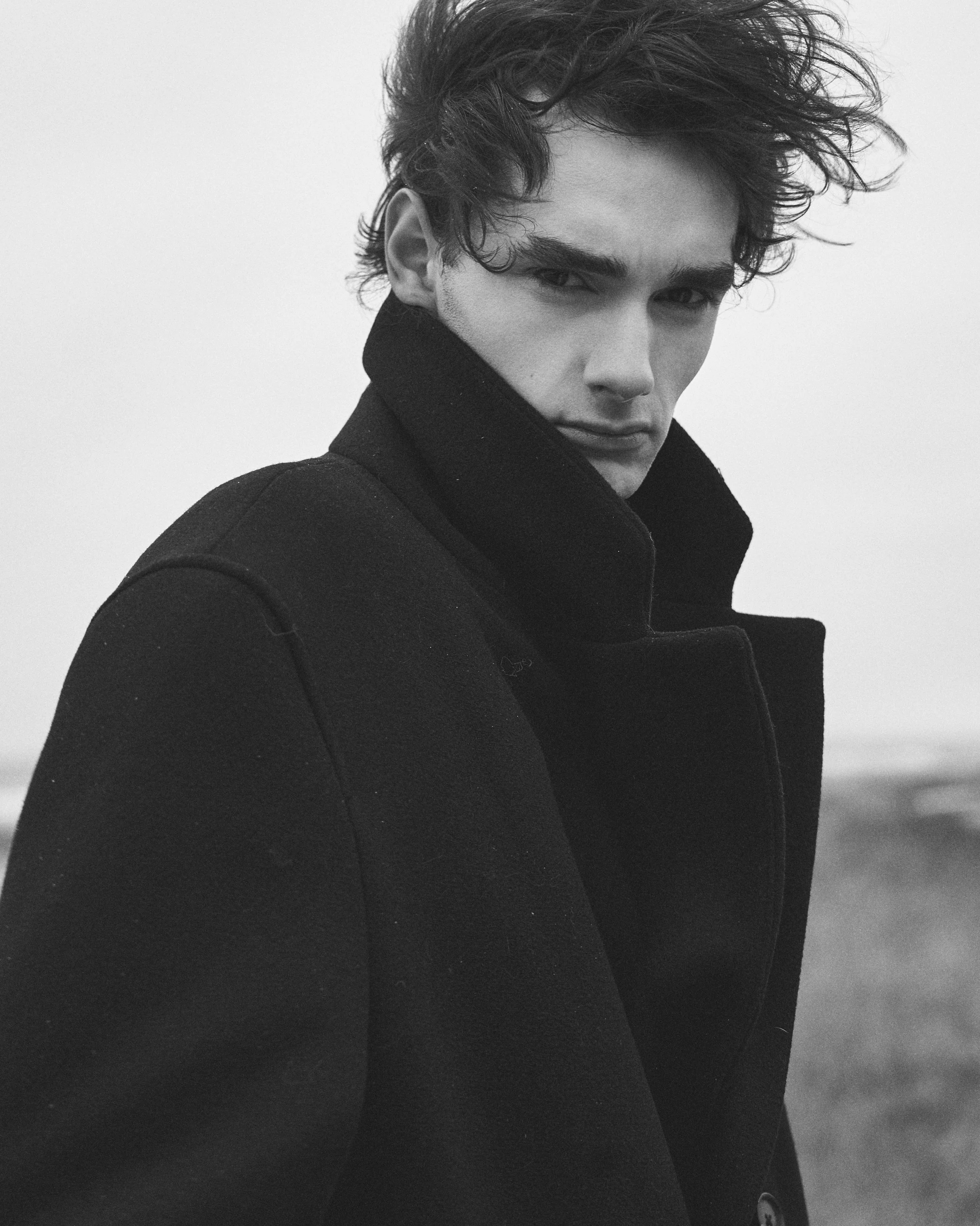 A black-and-white photo of a young man with tousled hair, wearing a dark coat with the collar turned up, looking directly at the camera with a serious expression.