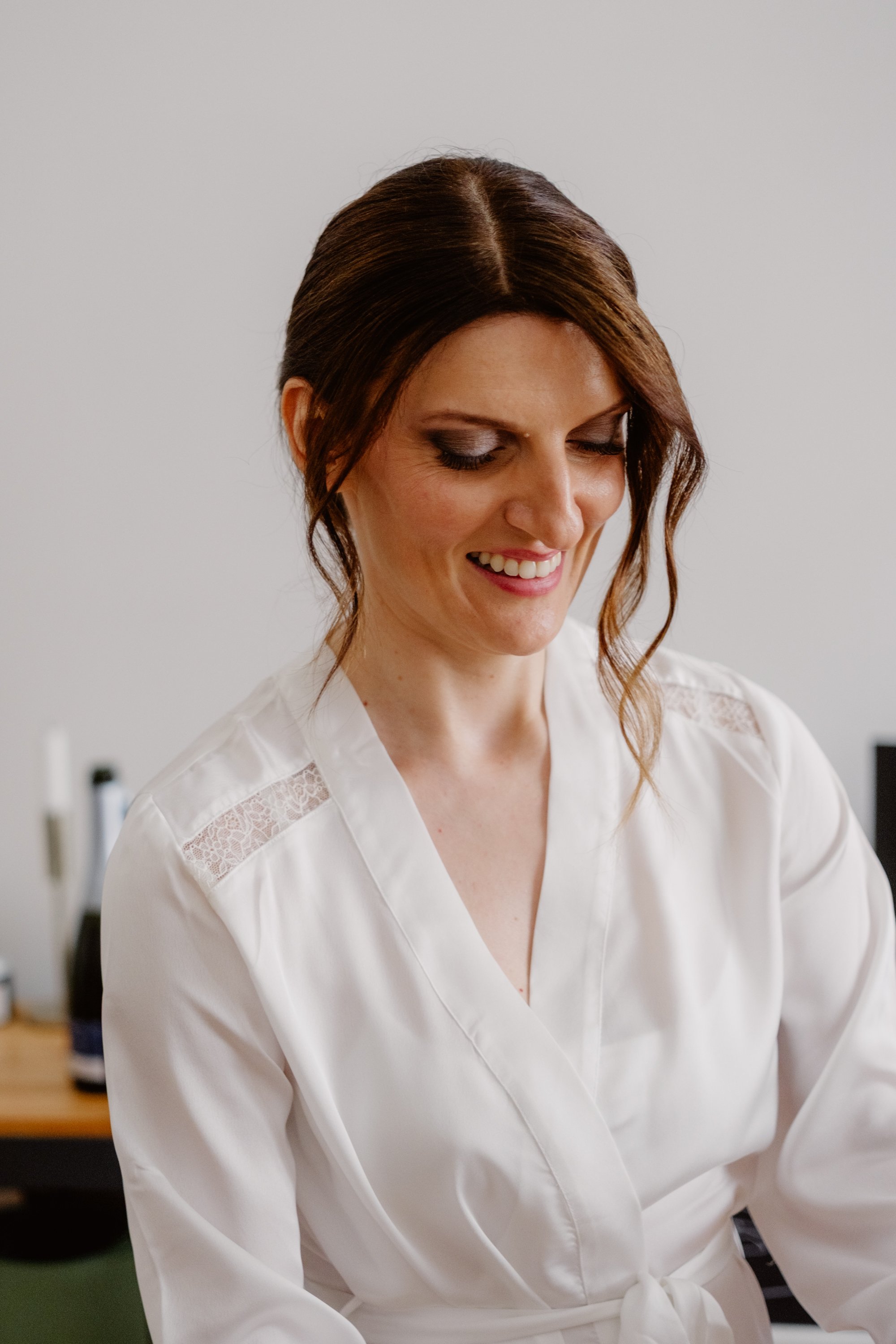 A bride with brown hair styled in loose waves, wearing a white robe with lace details, smiling and looking downward. The background is a plain, light-colored wall. It's her wedding day.