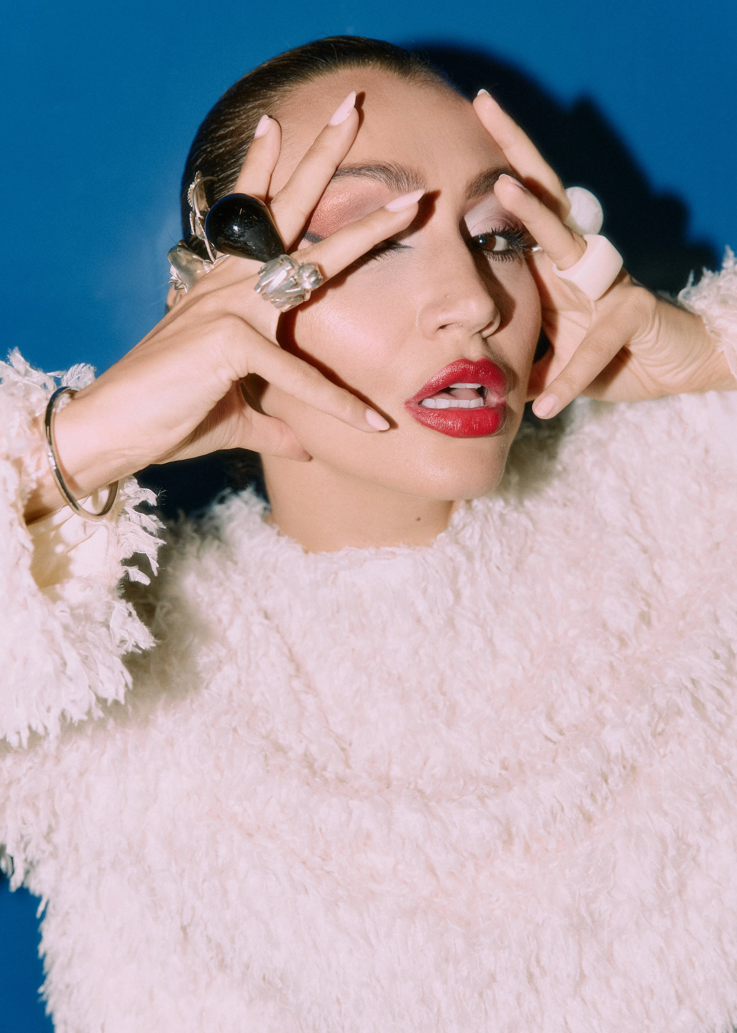 A woman with dark hair and makeup poses with her hands framing her face, wearing rings and accessories, against a blue background and a pink textured surface.