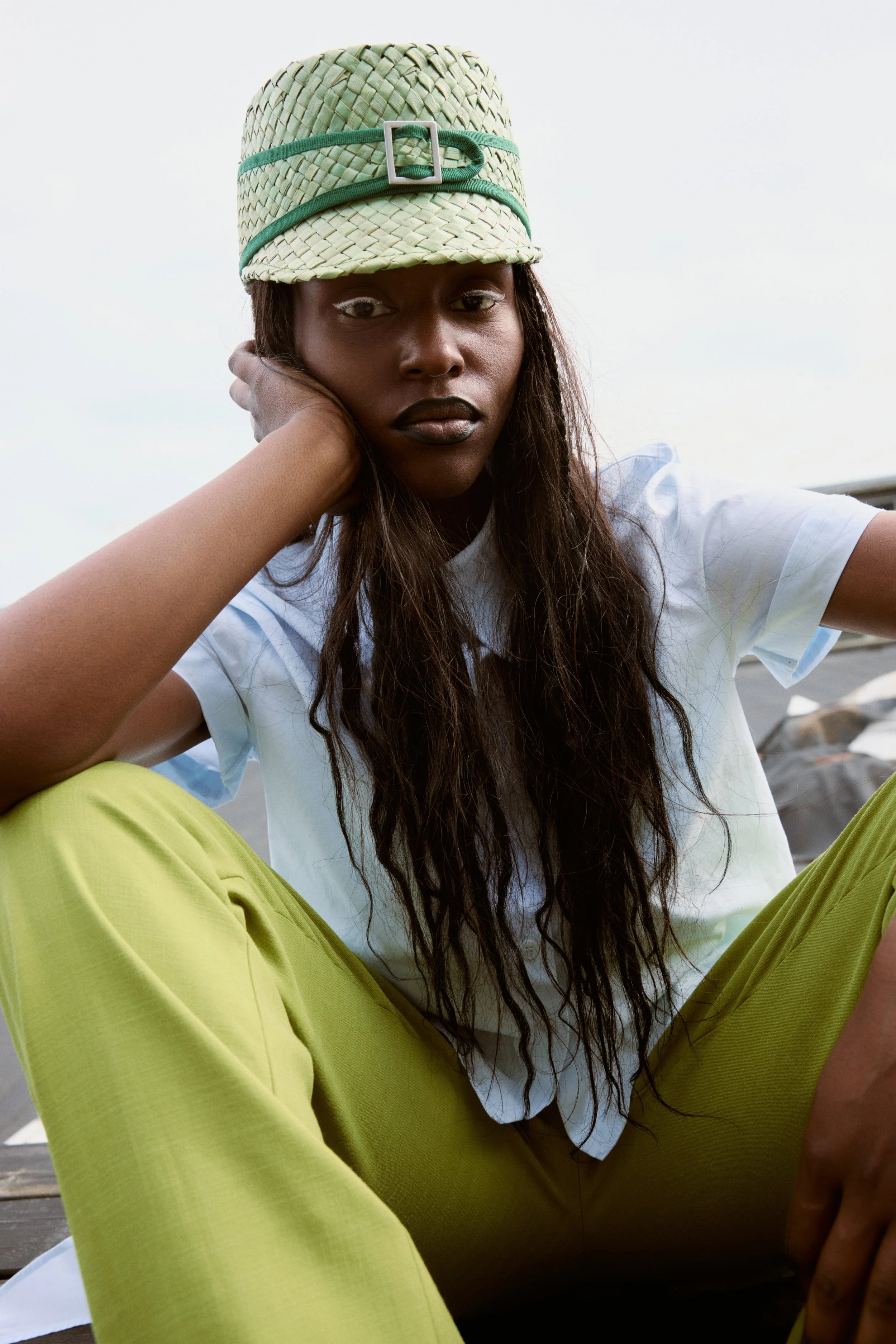 A woman with long, curly hair wearing a straw hat, a white T-shirt, and bright yellow pants, sitting outdoors with a cloudy sky in the background.