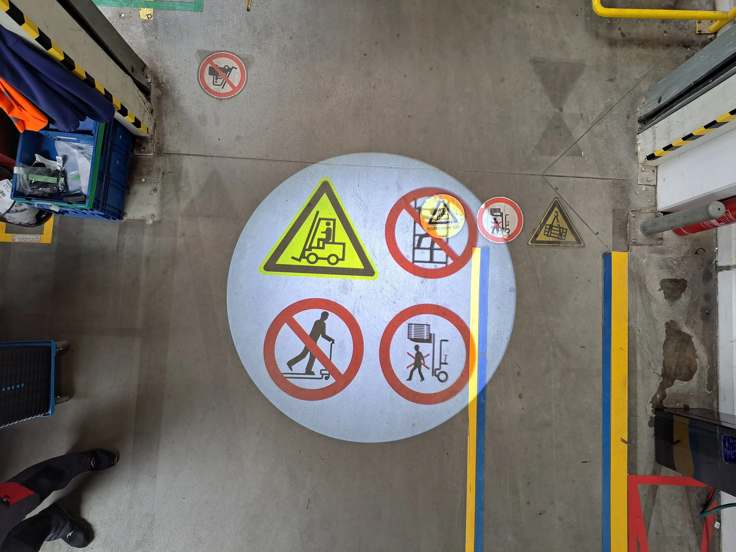 Projected safety and warning signs on the floor of a parking garage indicating no climbing, no walking on trolleys, no lifting heavy objects, and caution for forklift operation.
