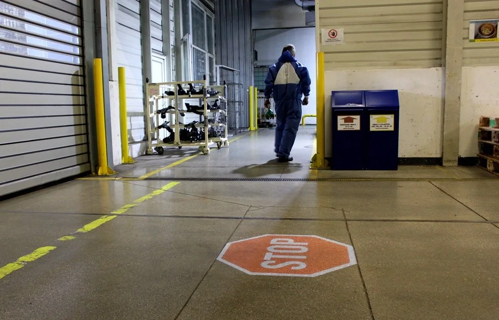 Warehouse aisle with a stop sign projected on the floor, a person walking away, yellow safety barriers, and a blue recycling bin.
