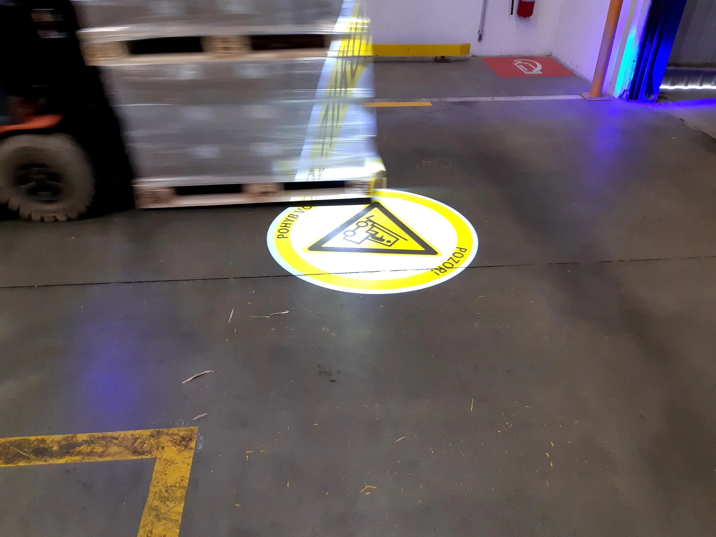 Warehouse floor with a yellow and black safety sign indicating caution, and a forklift moving pallets.