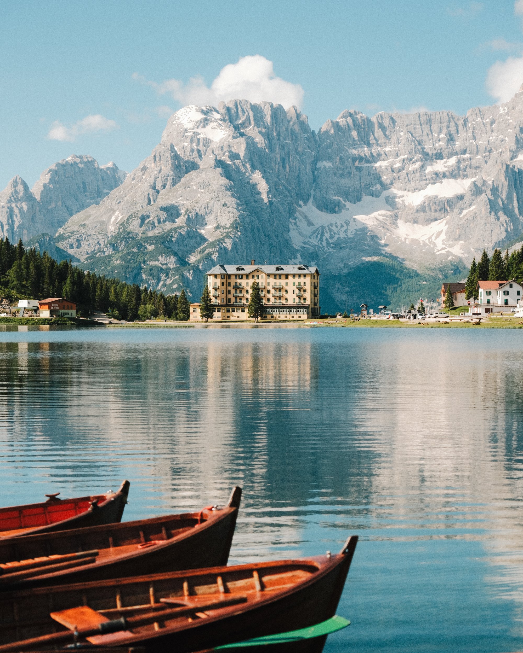 Bateaux en bois sur un lac alpin avec montagnes enneigées et bâtiments résidentiels en arrière-plan, sous un ciel bleu avec quelques nuages. Paysages, photo, montagne, rando, randonnée, sport, outdoor, exposition. Misurina, dolomites, italie