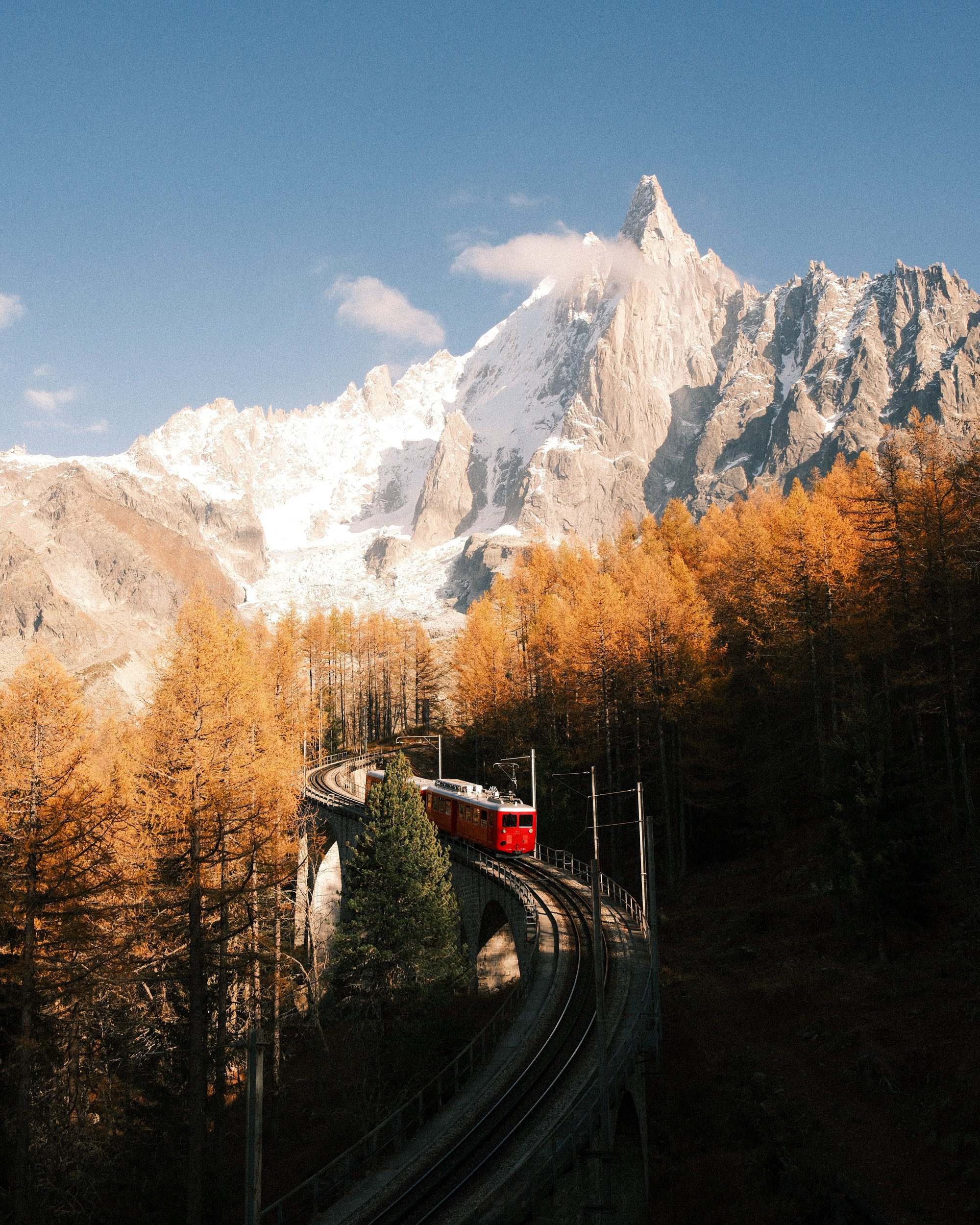 Train rouge voyageant sur un viaduc à travers une forêt automnale avec des montagnes enneigées en arrière-plan. Paysages, photo, montagne, rando, randonnée, sport, outdoor, exposition, signal forbes, Chamonix, les drus