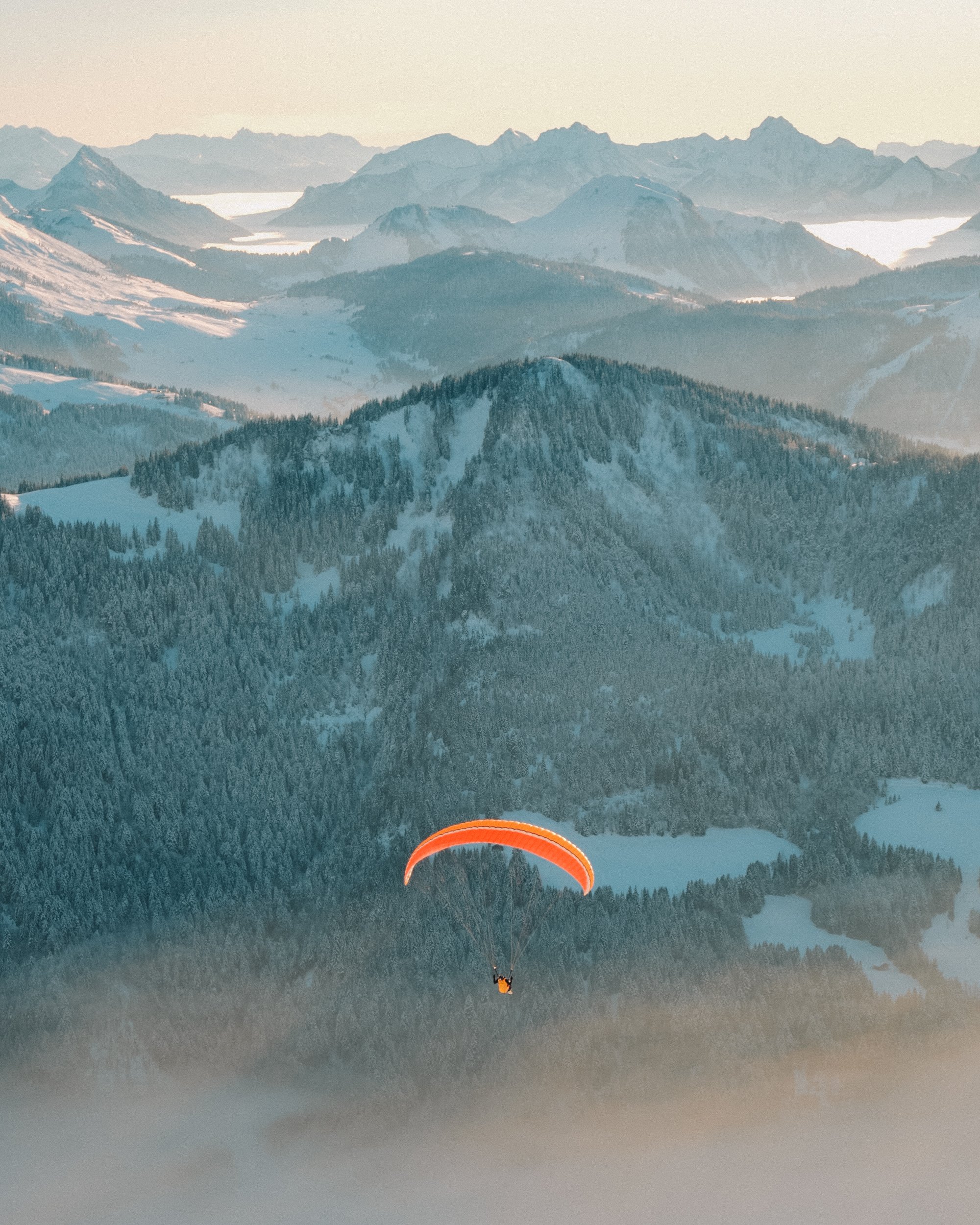Parapente orange volant au-dessus d'une forêt enneigée dans une vallée montagneuse. Paysages, photo, montagne, rando, randonnée, sport, outdoor, exposition