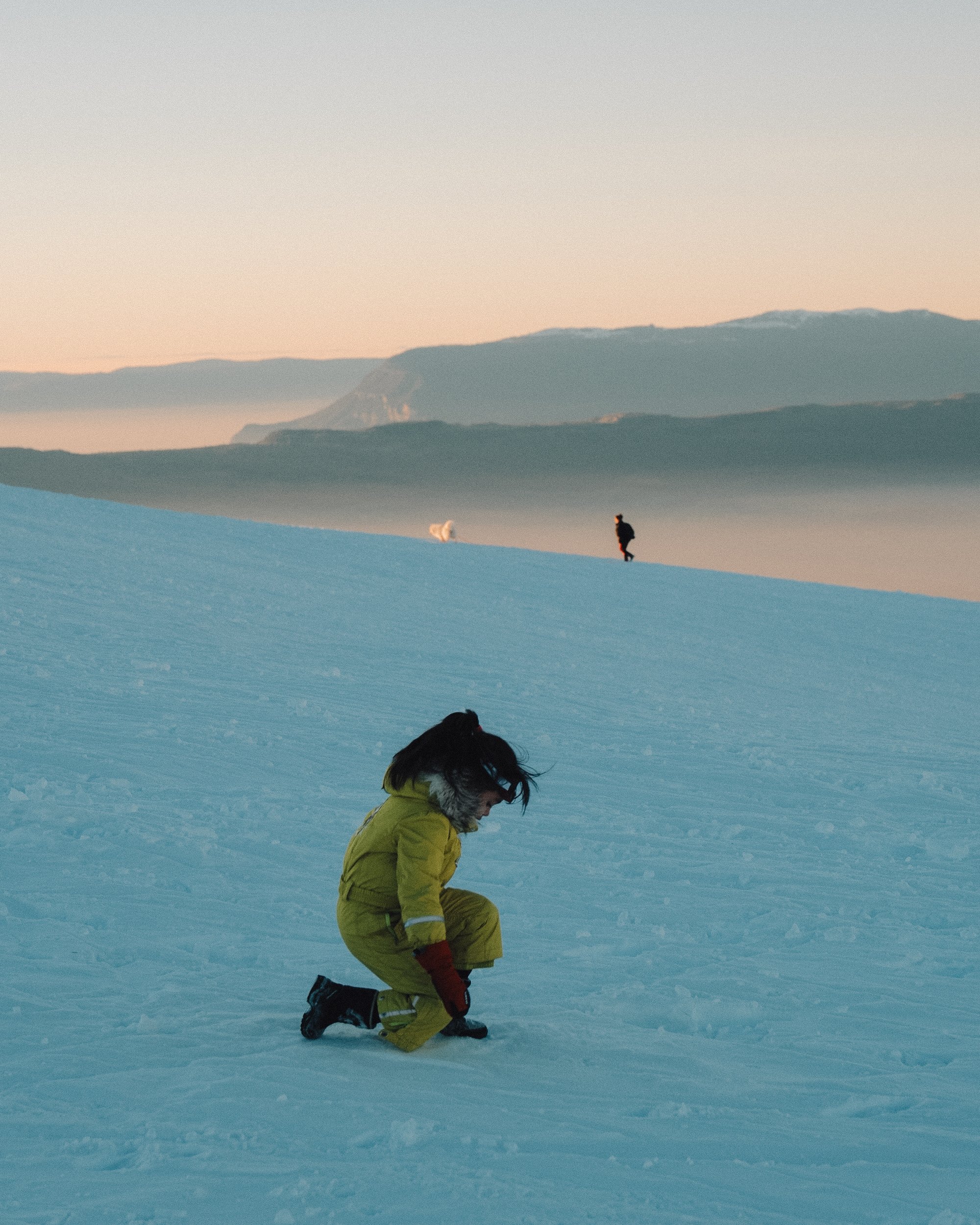 Une femme en combinaison jaune ramasse la neige sur un paysage enneigé avec deux autres personnes en arrière-plan lors d'un coucher de soleil. Paysages, photo, montagne, rando, randonnée, sport, outdoor, exposition