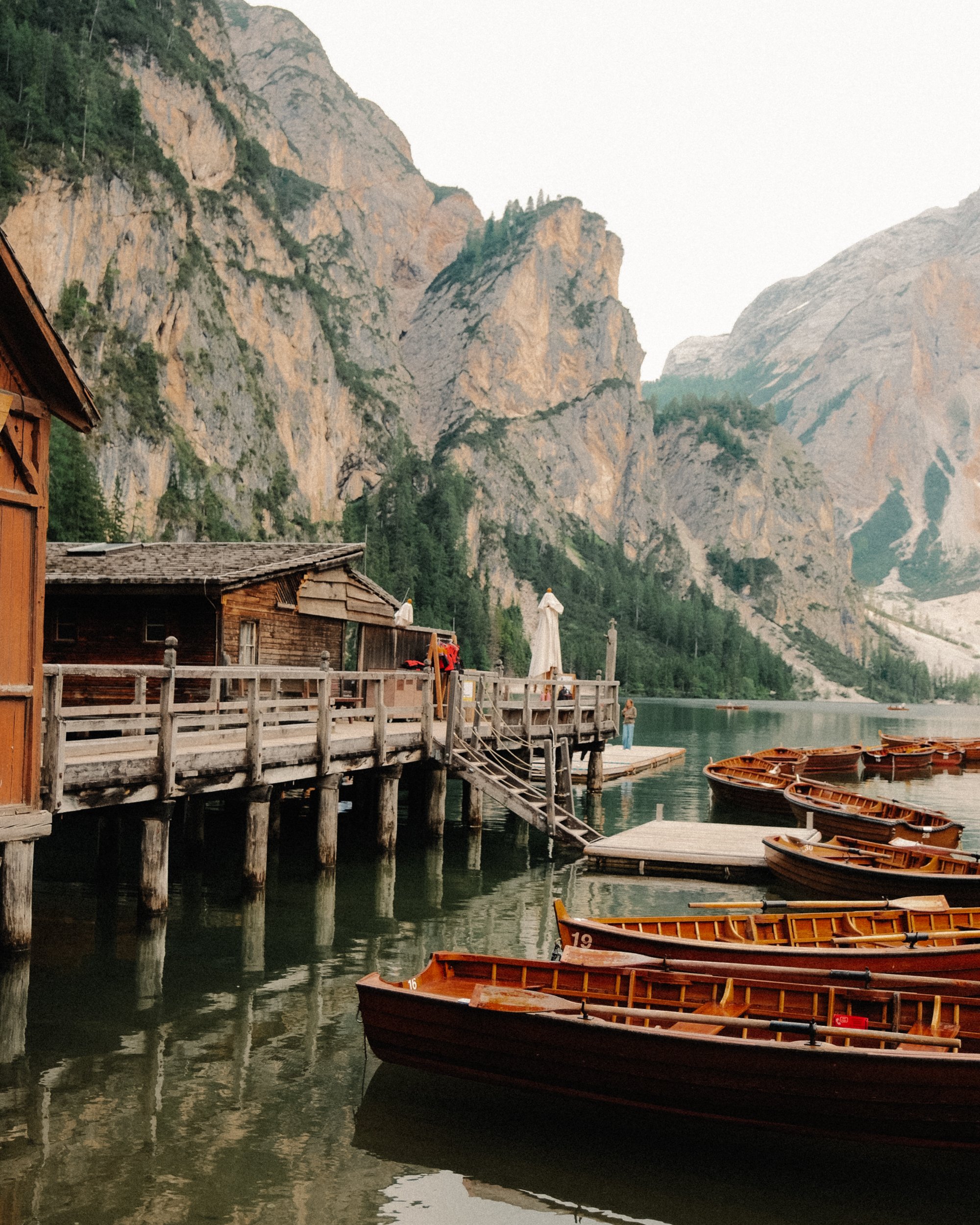 Un quai en bois avec plusieurs bateaux en bois amarrés sur un lac entouré de montagnes rocheuses et de forêts, avec une petite cabane en bois sur le quai. Paysages, photo, montagne, rando, randonnée, sport, outdoor, exposition. Dolomites